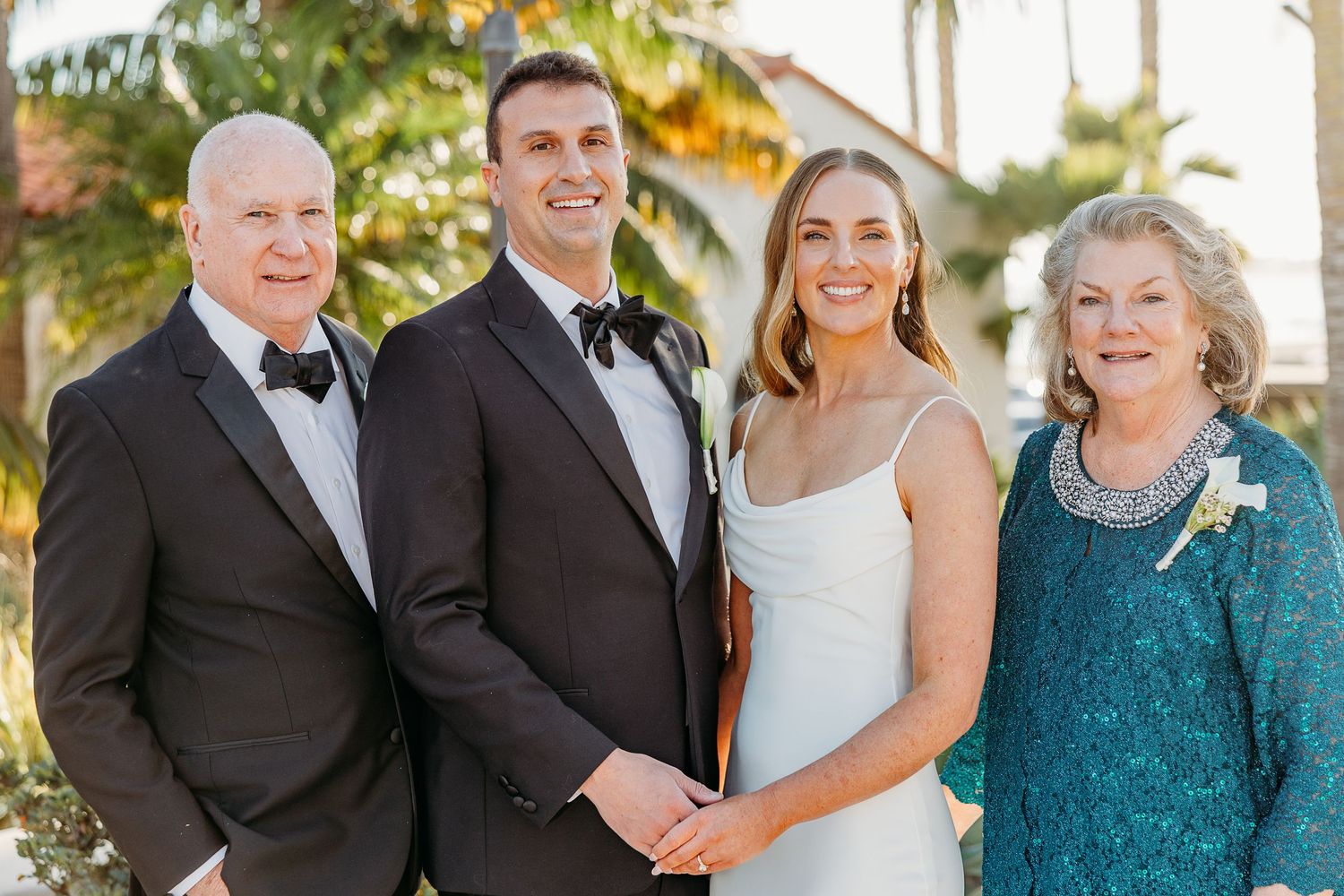 Wedding party gathered outdoors in formal attire with teal and black color scheme against a sunny backdrop.