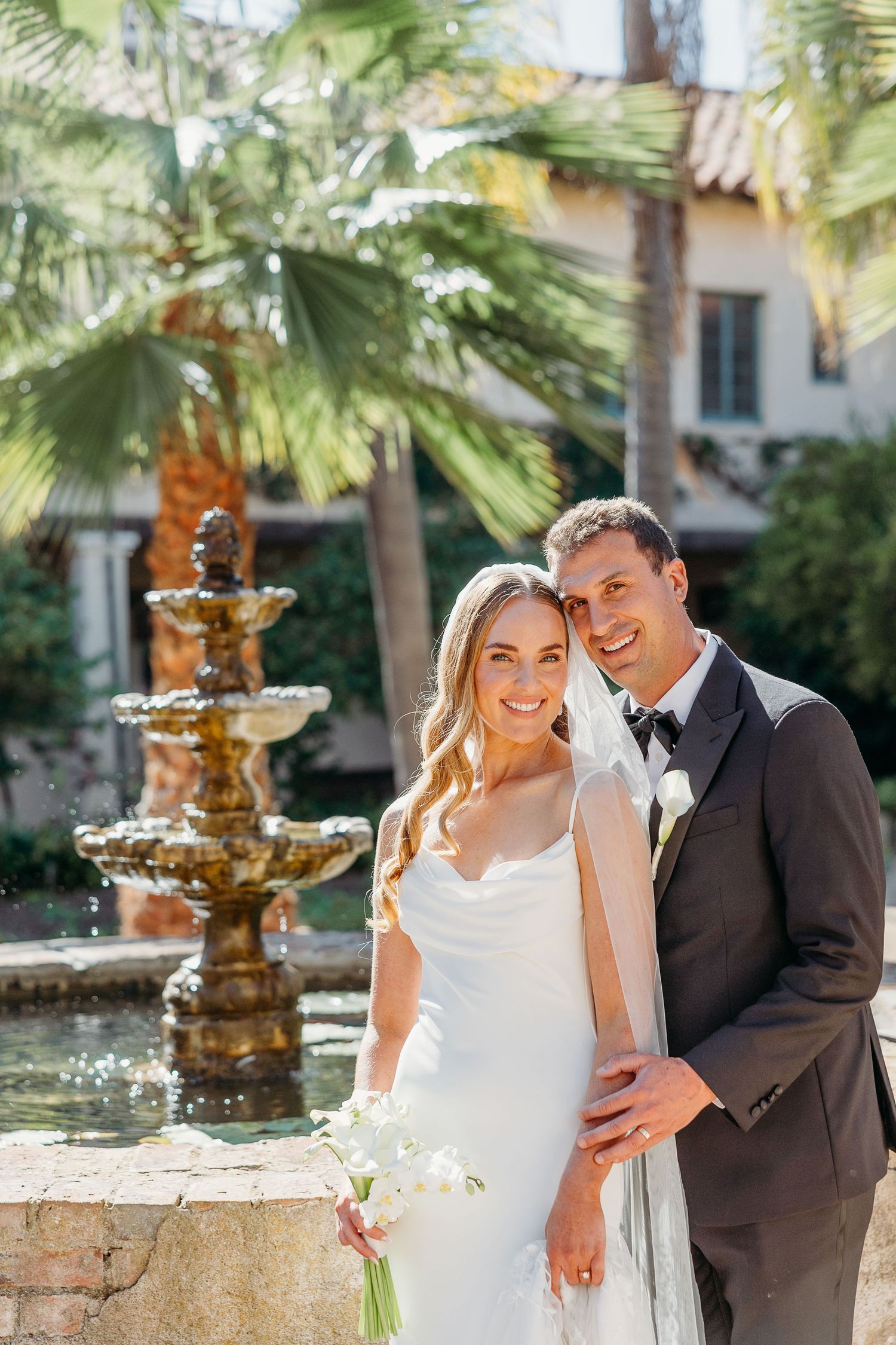 A romantic wedding photo featuring a couple posing by an ornate fountain with palm trees in a Mediterranean-style courtyard.