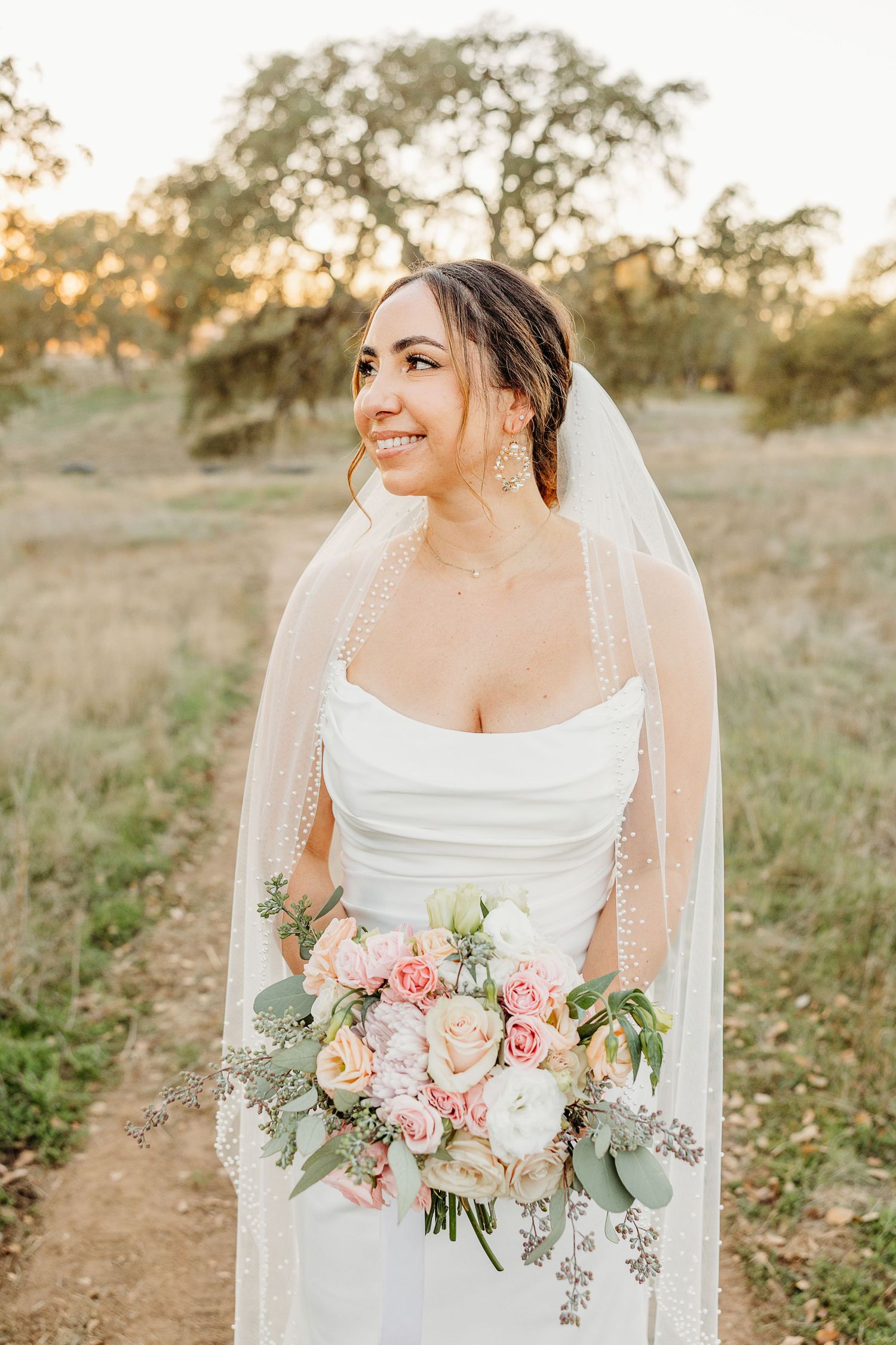 Bride in elegant white dress holding pastel bouquet in outdoor setting at sunset.