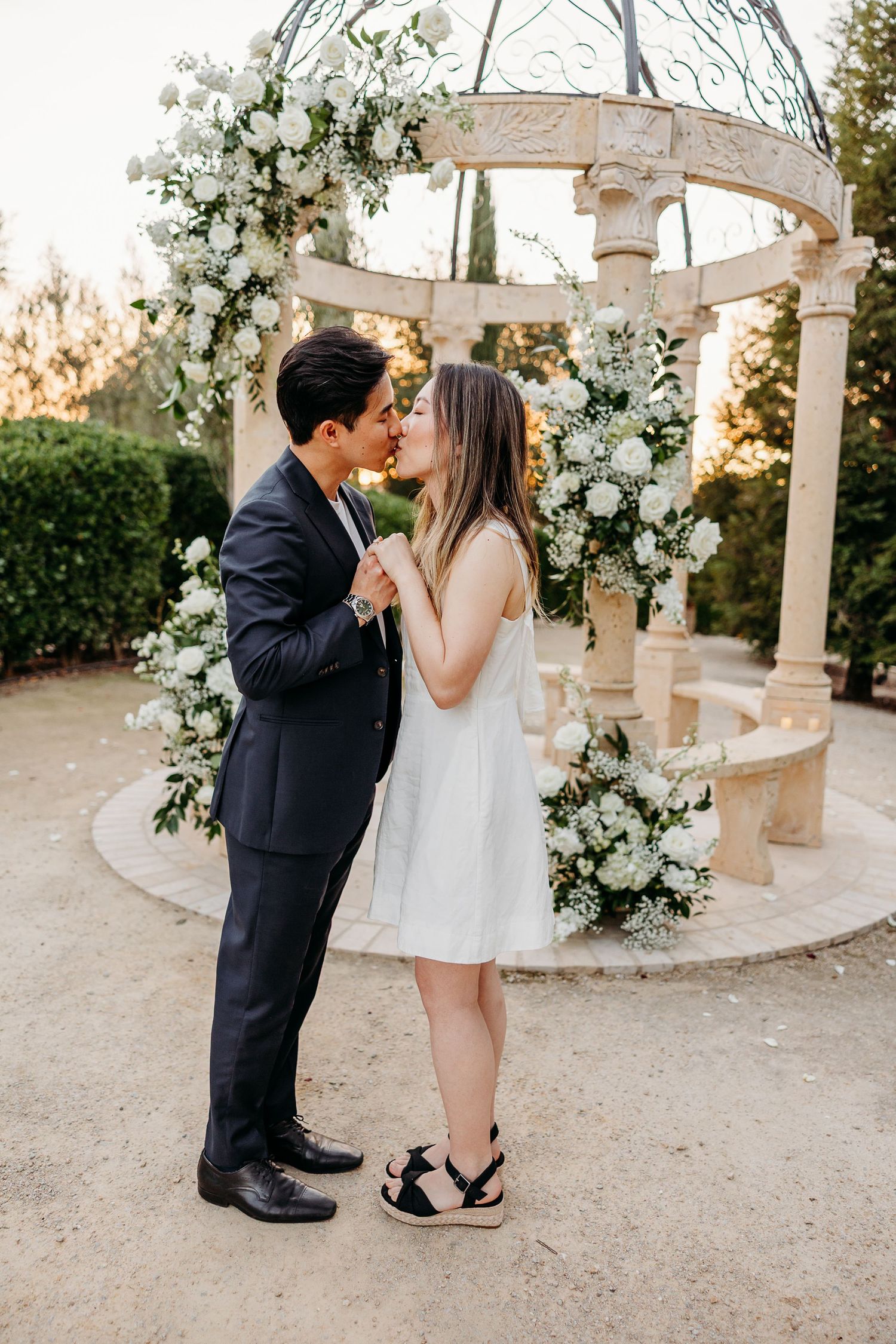 A tender moment between a couple beneath an ornate gazebo adorned with cascading white flowers with setting sun golden light in the
