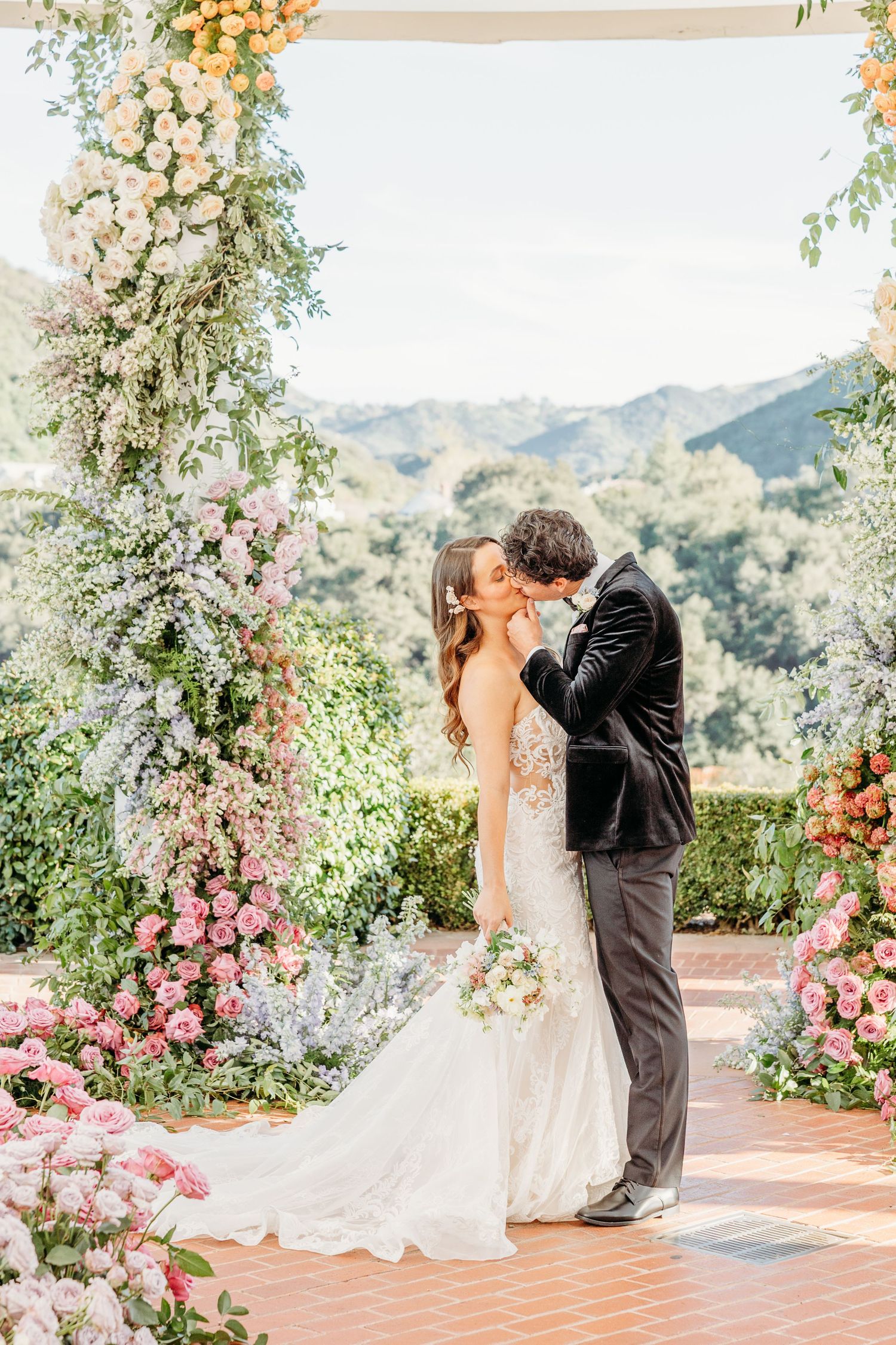A romantic wedding ceremony under a floral arch with mountain views, featuring pink and white roses and greenery decorations.