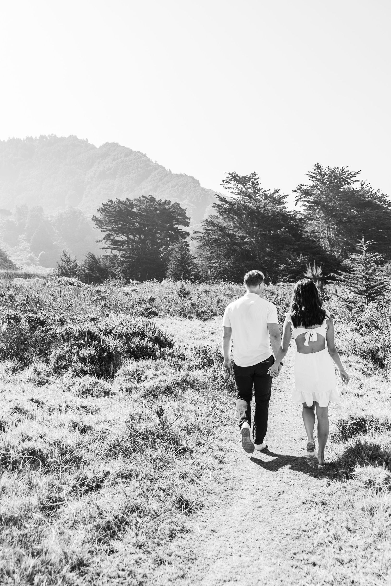 Black and white photo of a couple walking hand in hand on a scenic mountain path.