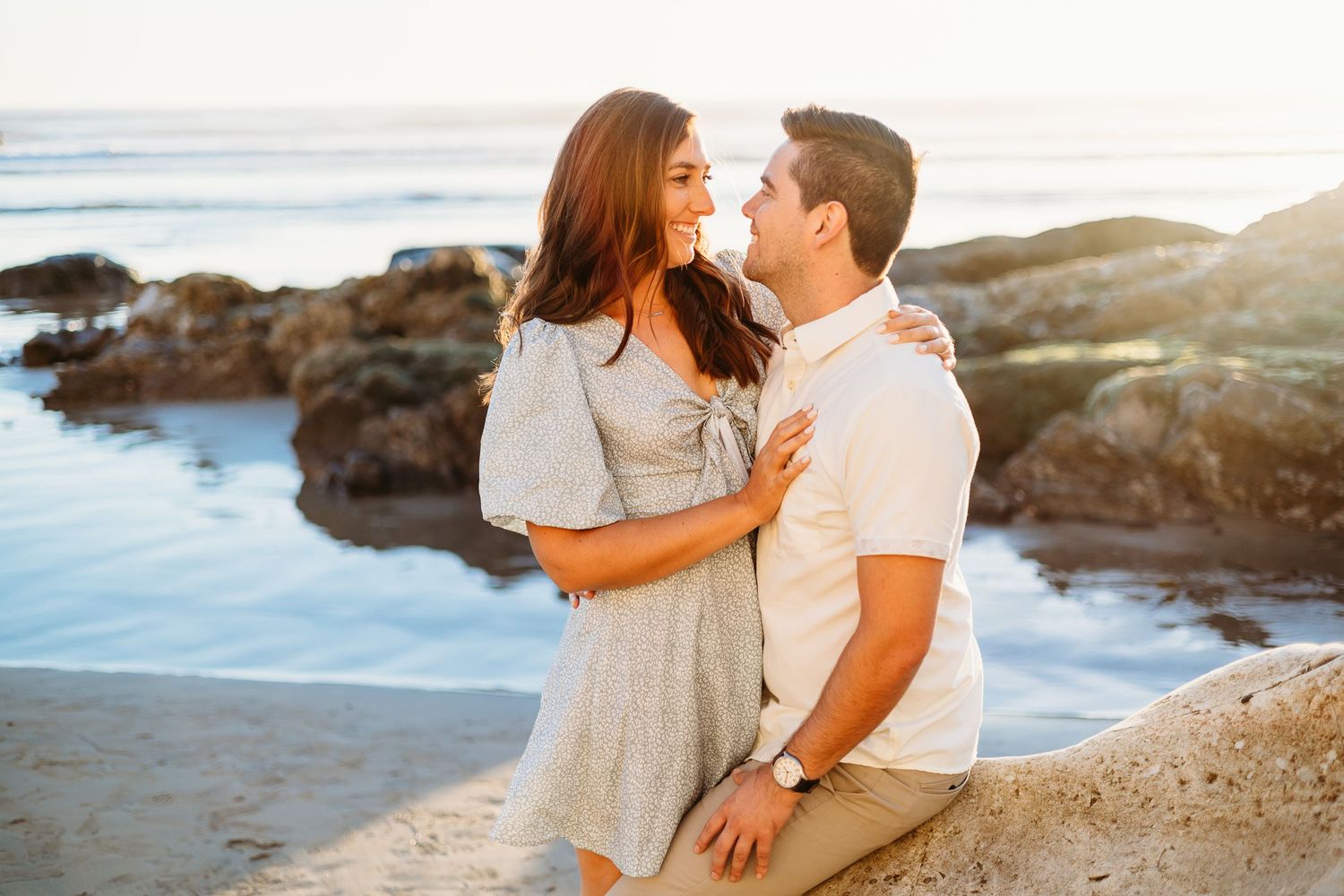A couple shares an intimate moment on coastal rocks with ocean waves behind them.
