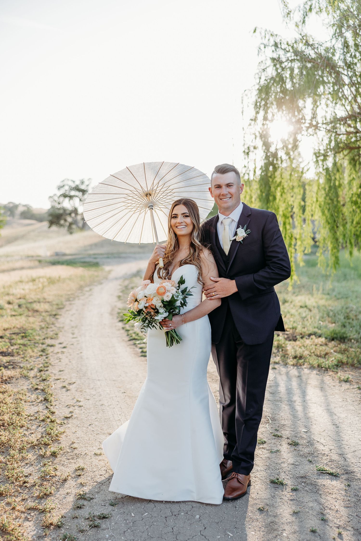 Romantic wedding couple with parasol poses at sunset on a rustic dirt path surrounded by willow trees.