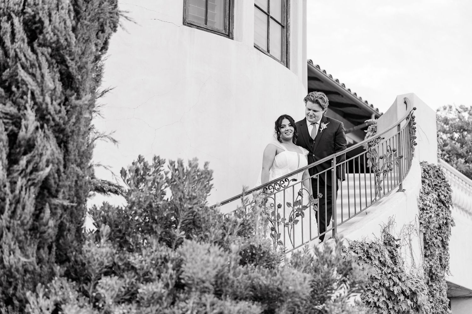 A couple stands on a Mediterranean-style balcony with white stucco walls and wrought iron railings.