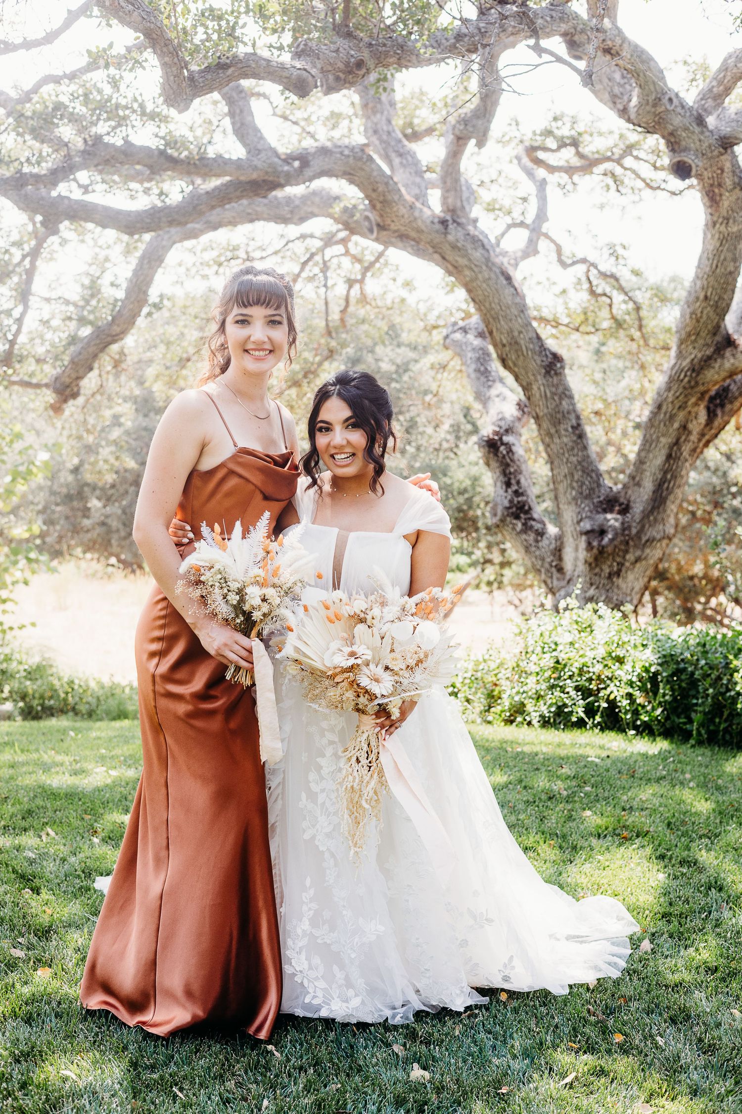 Bridesmaid in rust-colored silk dress poses with bride in lace gown under sprawling oak trees.