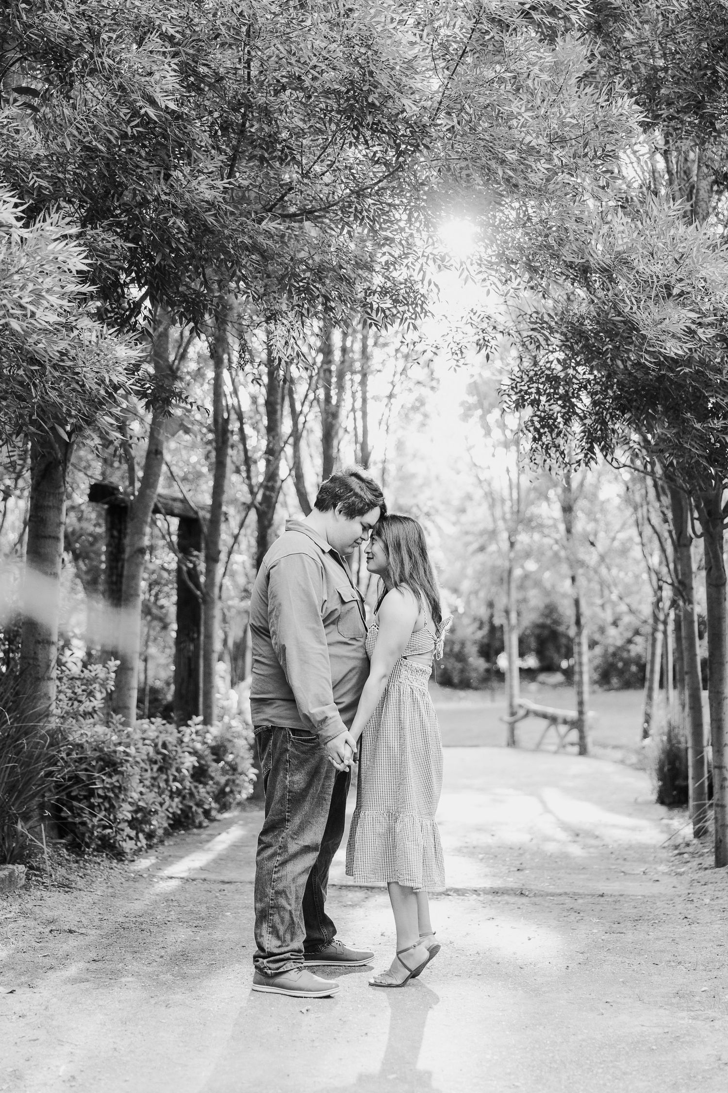 A romantic couple stands close together on a tree-lined path in black and white.