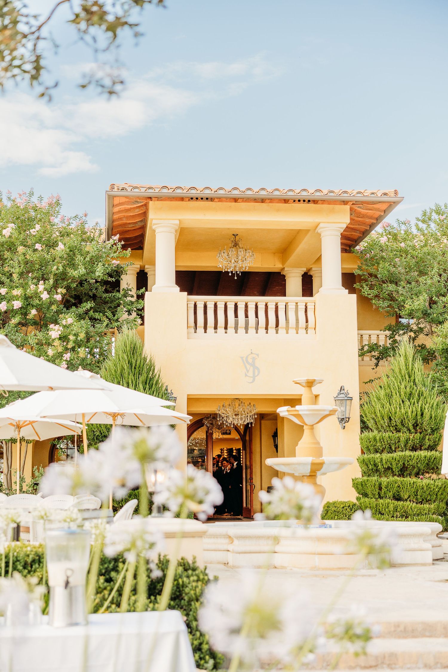 Mediterranean style villa with cream stucco exterior, columns, and balcony surrounded by lush landscaping and white umbrellas.