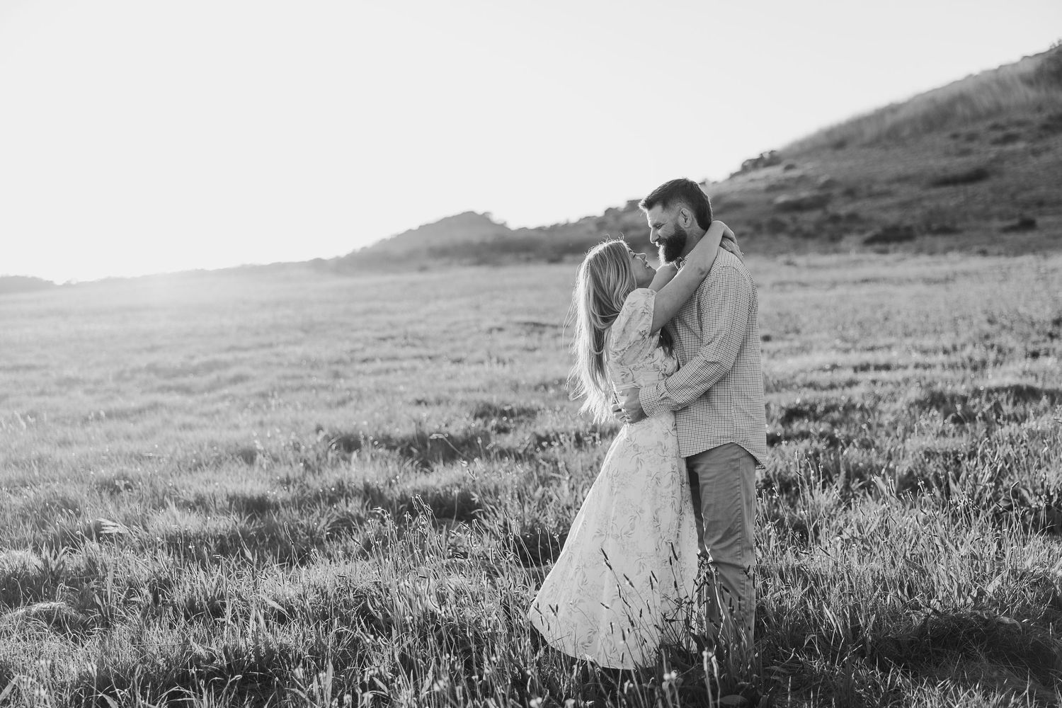 A romantic black and white photograph of a couple embracing in a sunlit meadow with rolling hills in the background.