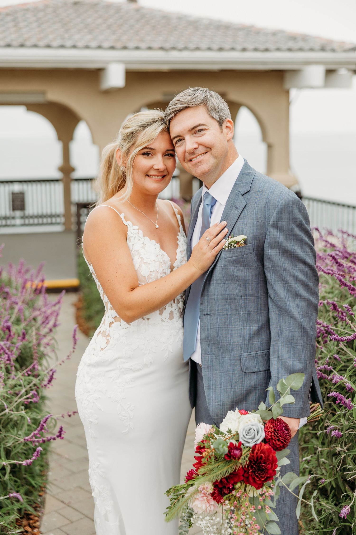 Romantic wedding portrait against coastal gazebo surrounded by purple flowering garden.