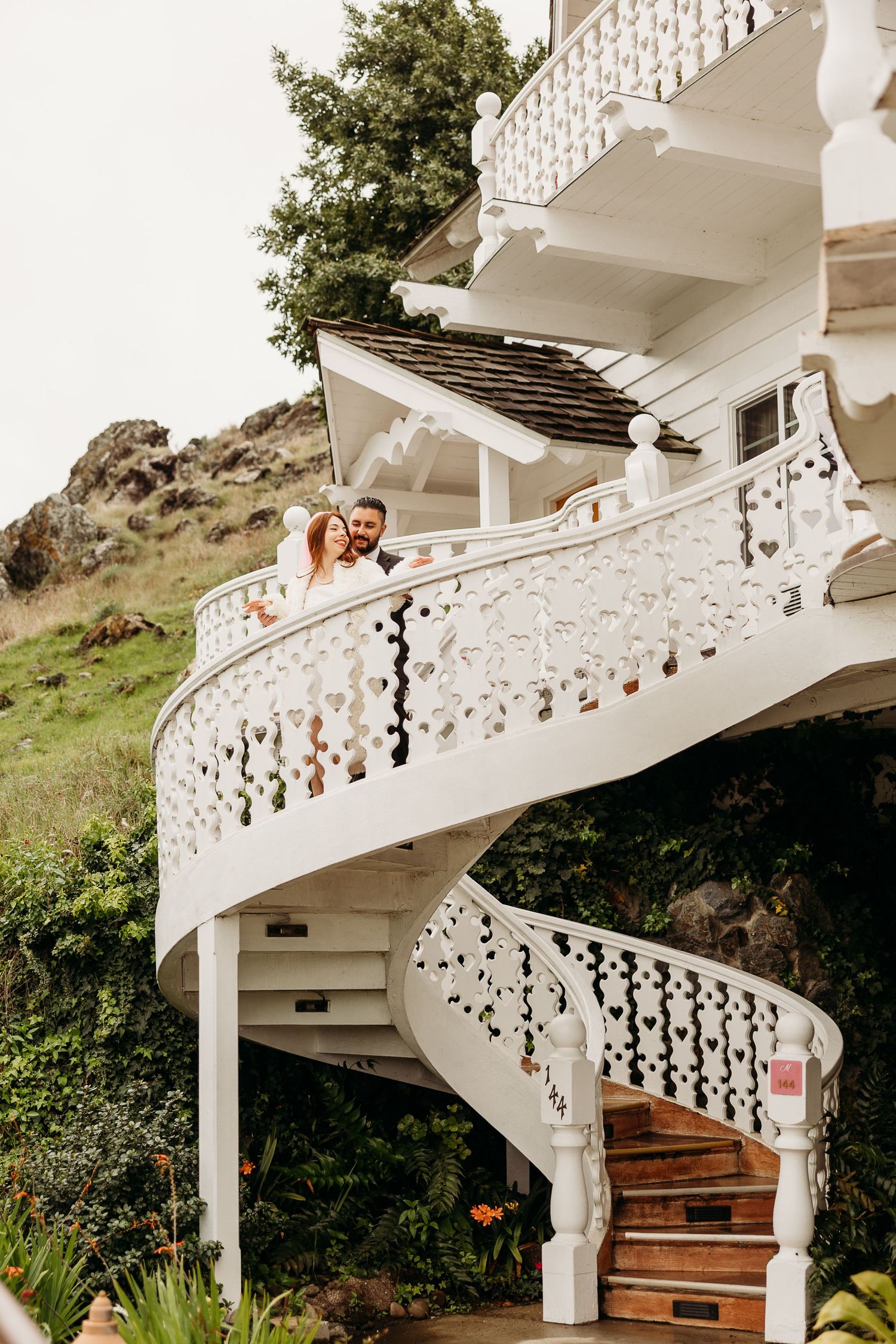 Rustic white staircase with decorative railings leading up to a balcony against a lush hillside background.
