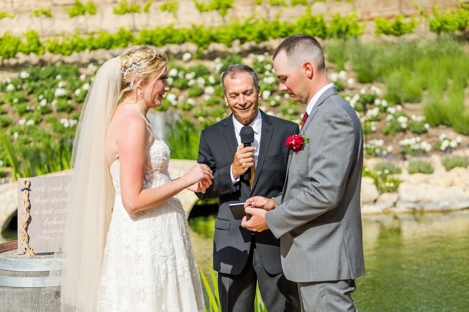 Intimate garden wedding ceremony featuring white lace dress and grey suit beside rustic wine barrels.