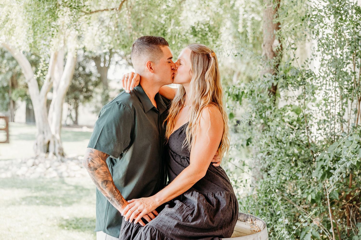 A romantic couple embraces outdoors surrounded by green trees and natural lighting.