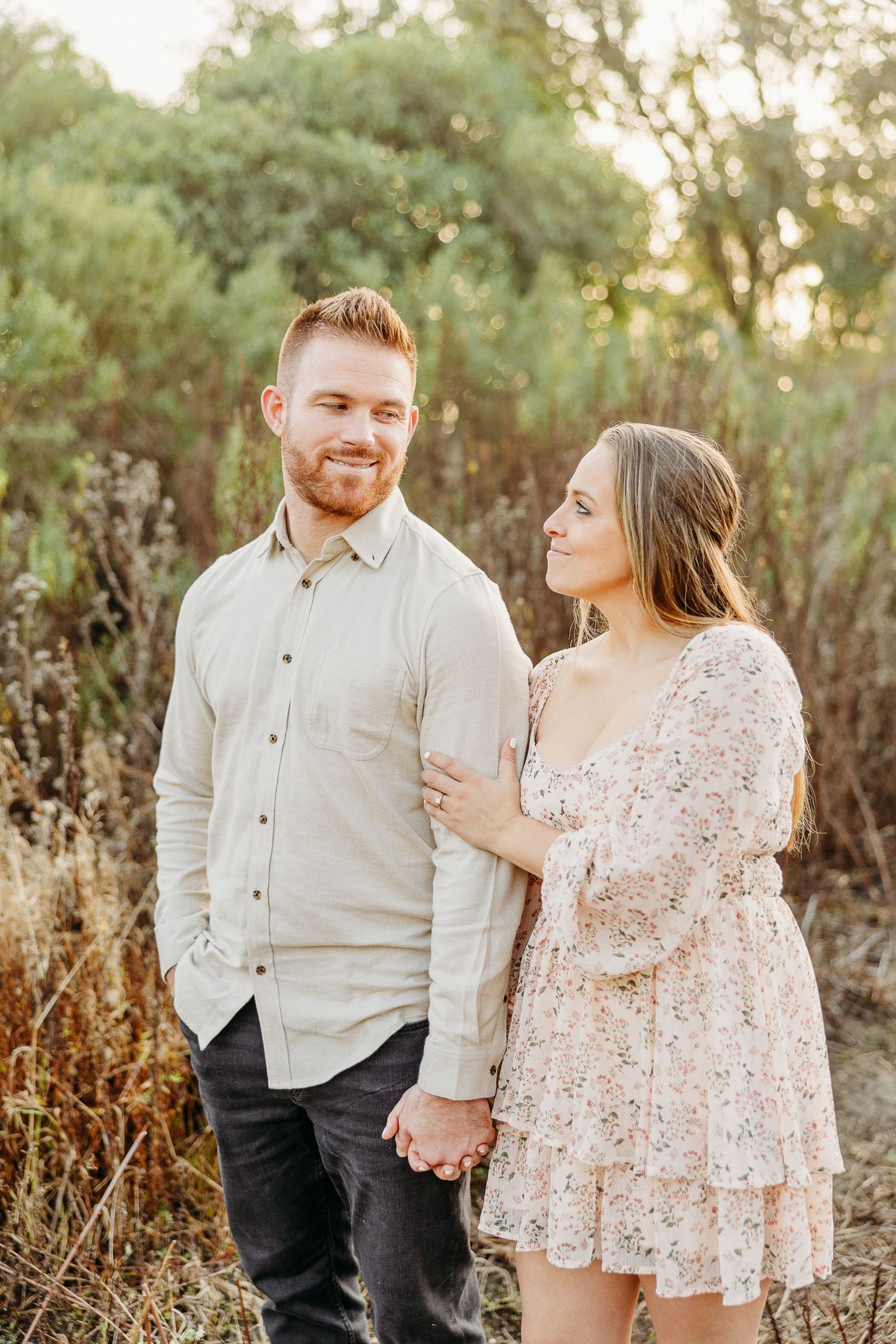Couple holding hands and smiling at each other in natural outdoor setting at sunset.