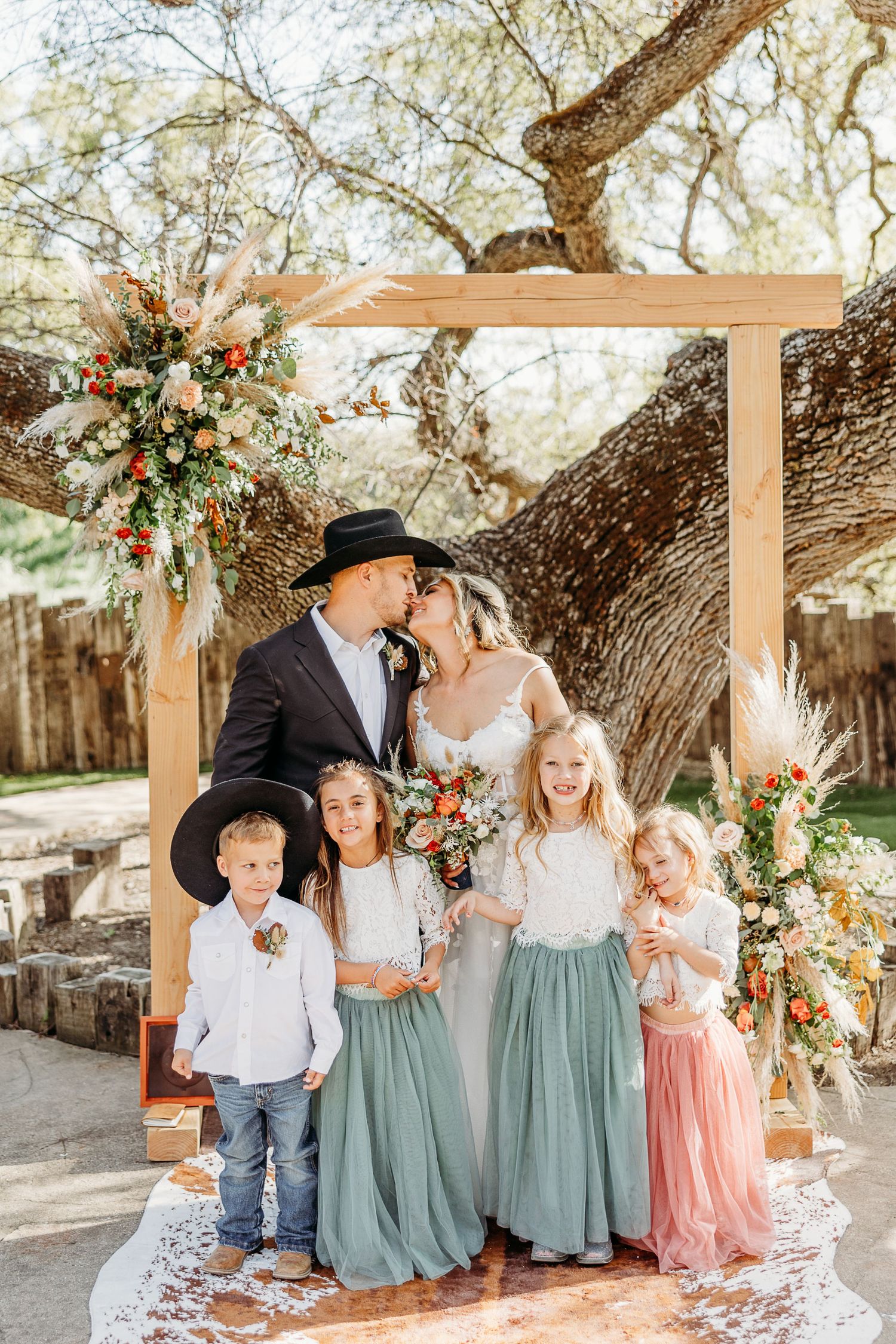 A western-themed outdoor wedding ceremony under a rustic wooden arch decorated with peach and white florals.