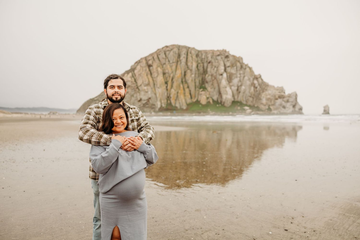 Couple sharing a tender moment on a beach with dramatic rock formations and reflective wet sand.