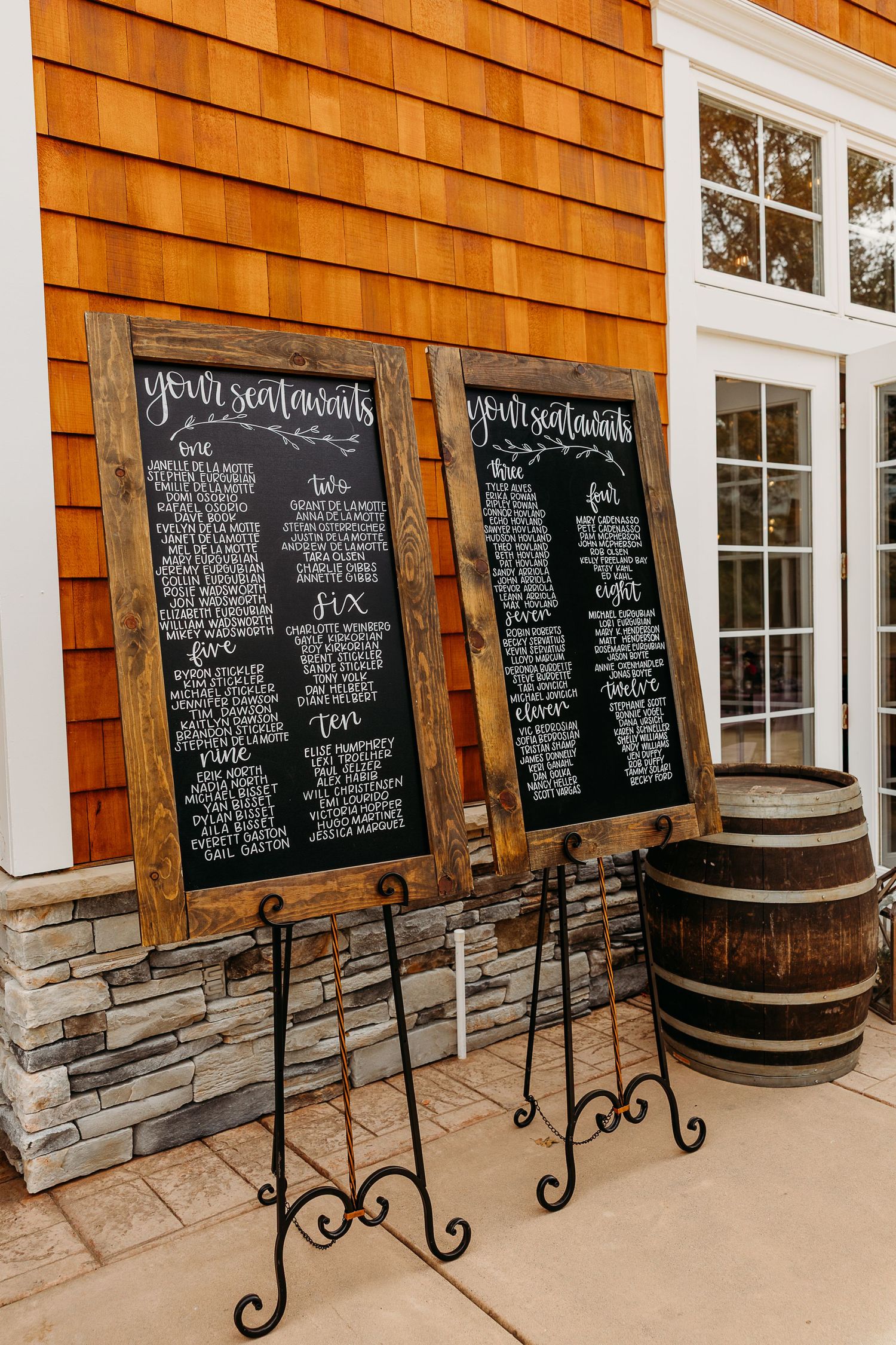 Wedding seating chart displayed on black chalkboards with wooden barrels and white trim exterior.