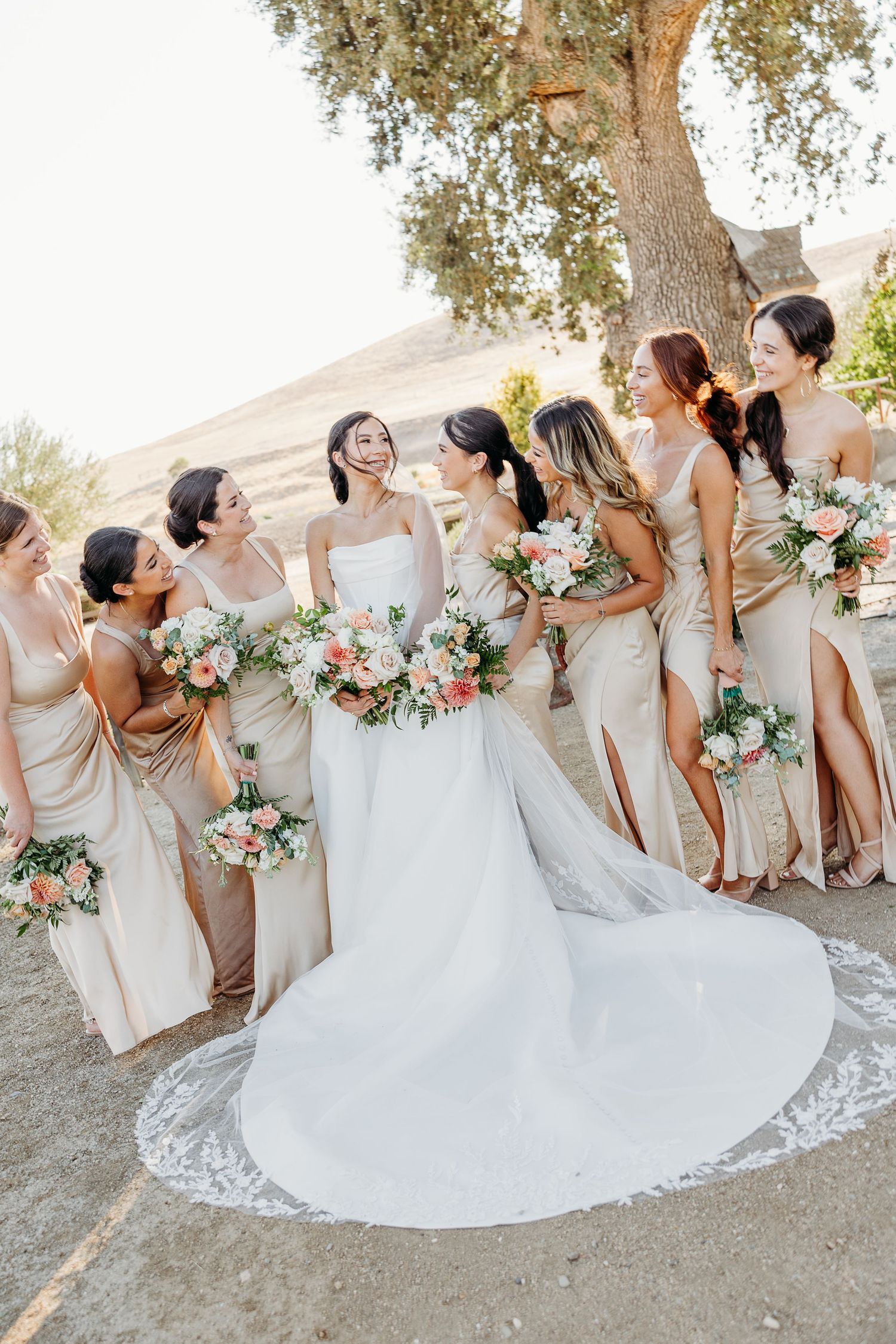 Bridesmaids in champagne dresses pose together with pink and white floral bouquets during an outdoor wedding celebration.