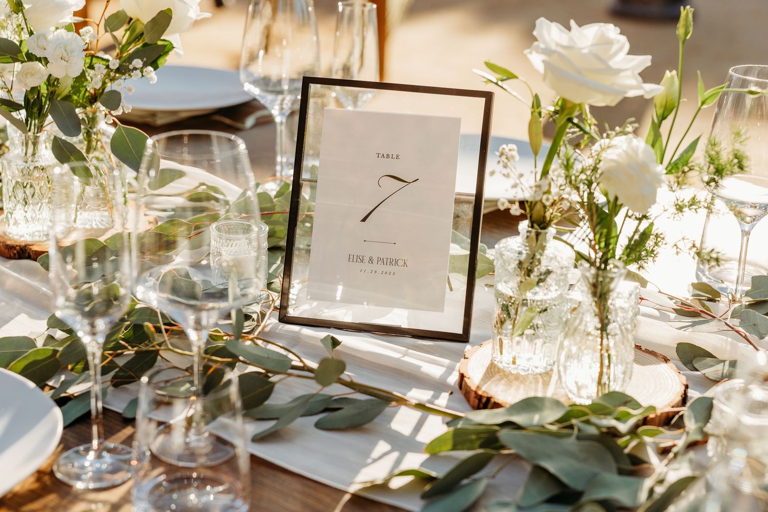 Elegant wedding table setting with framed table number, wine glasses, and eucalyptus leaf decorations in natural light.