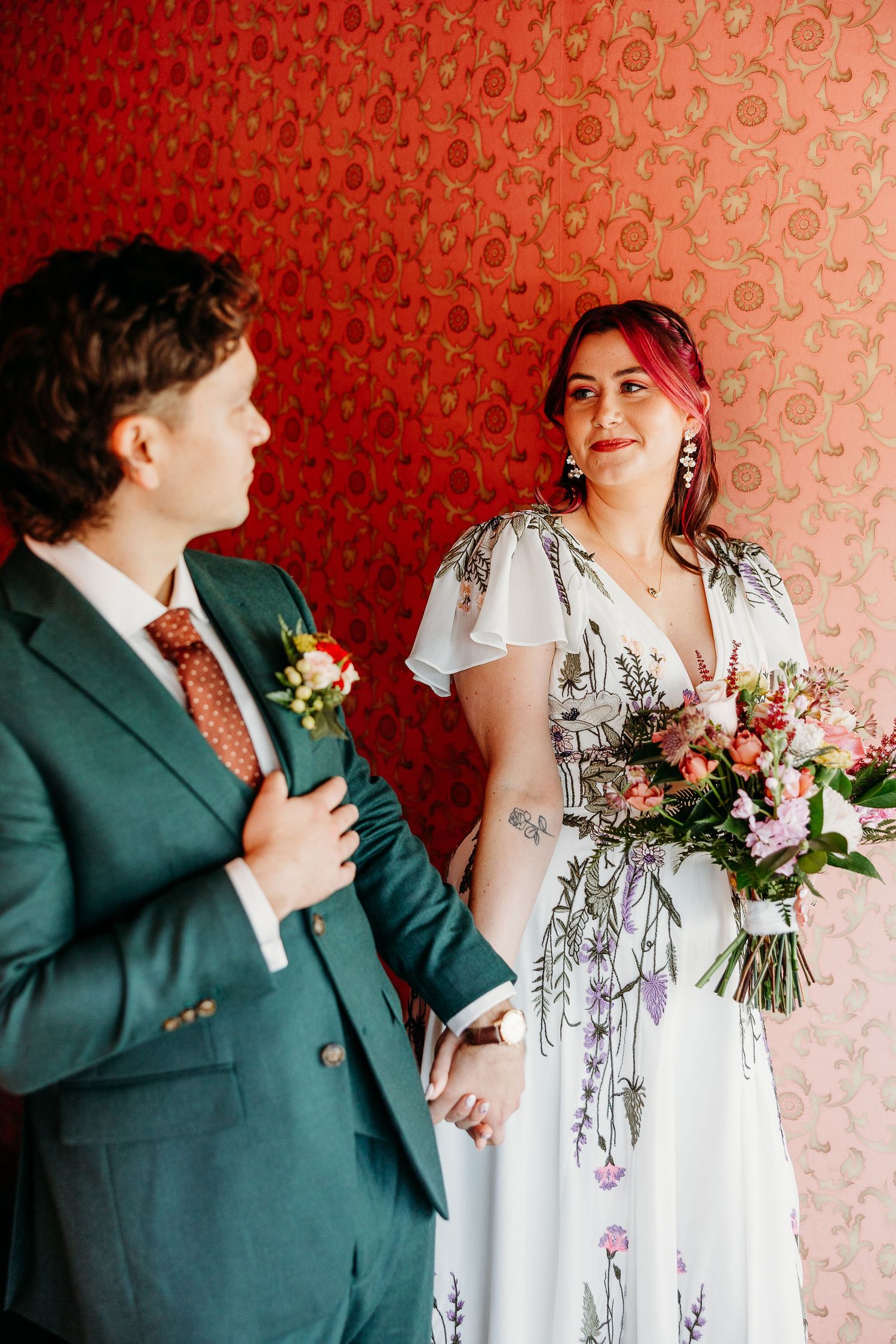 Wedding couple posing against red wallpaper with floral bouquet.