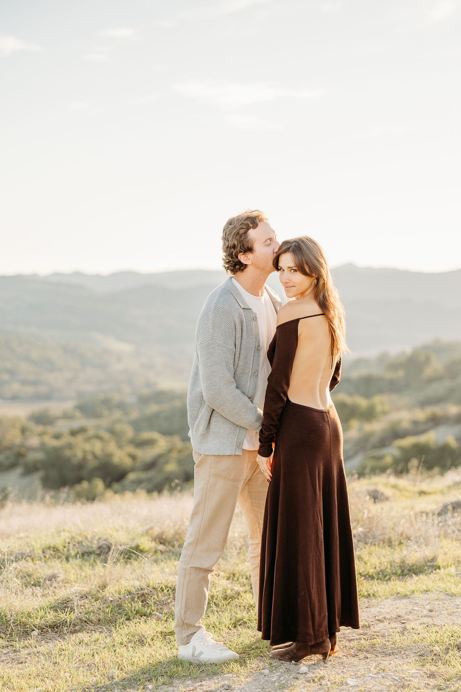 Couple shares romantic embrace on sunlit hillside during outdoor photoshoot.