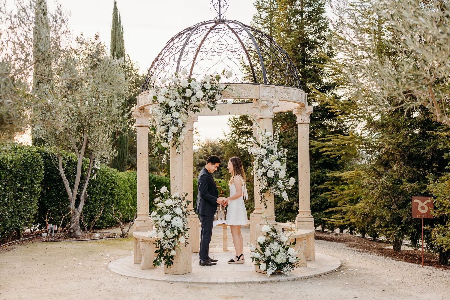 Couple stands under elegant gazebo with white floral arrangements at outdoor wedding ceremony.