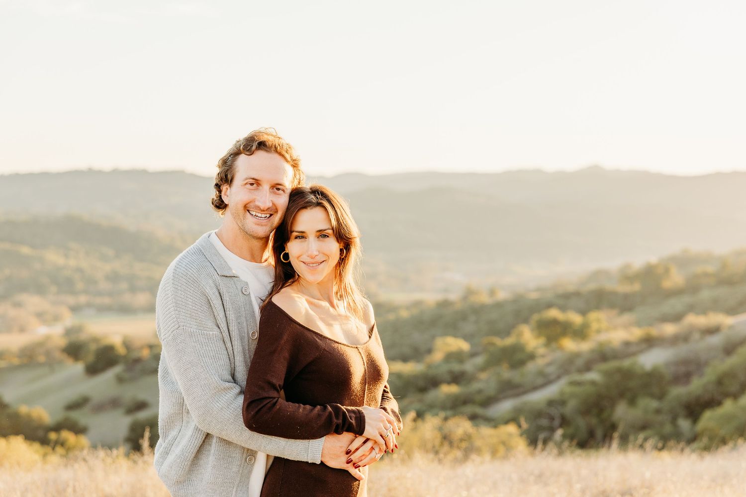 A couple embraces during golden hour with mountains and natural landscape behind them.
