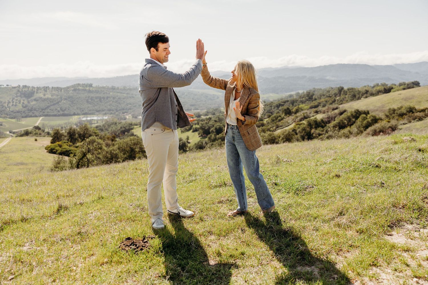 Two people give each other a high five while standing on a grassy hillside with mountains in the background.