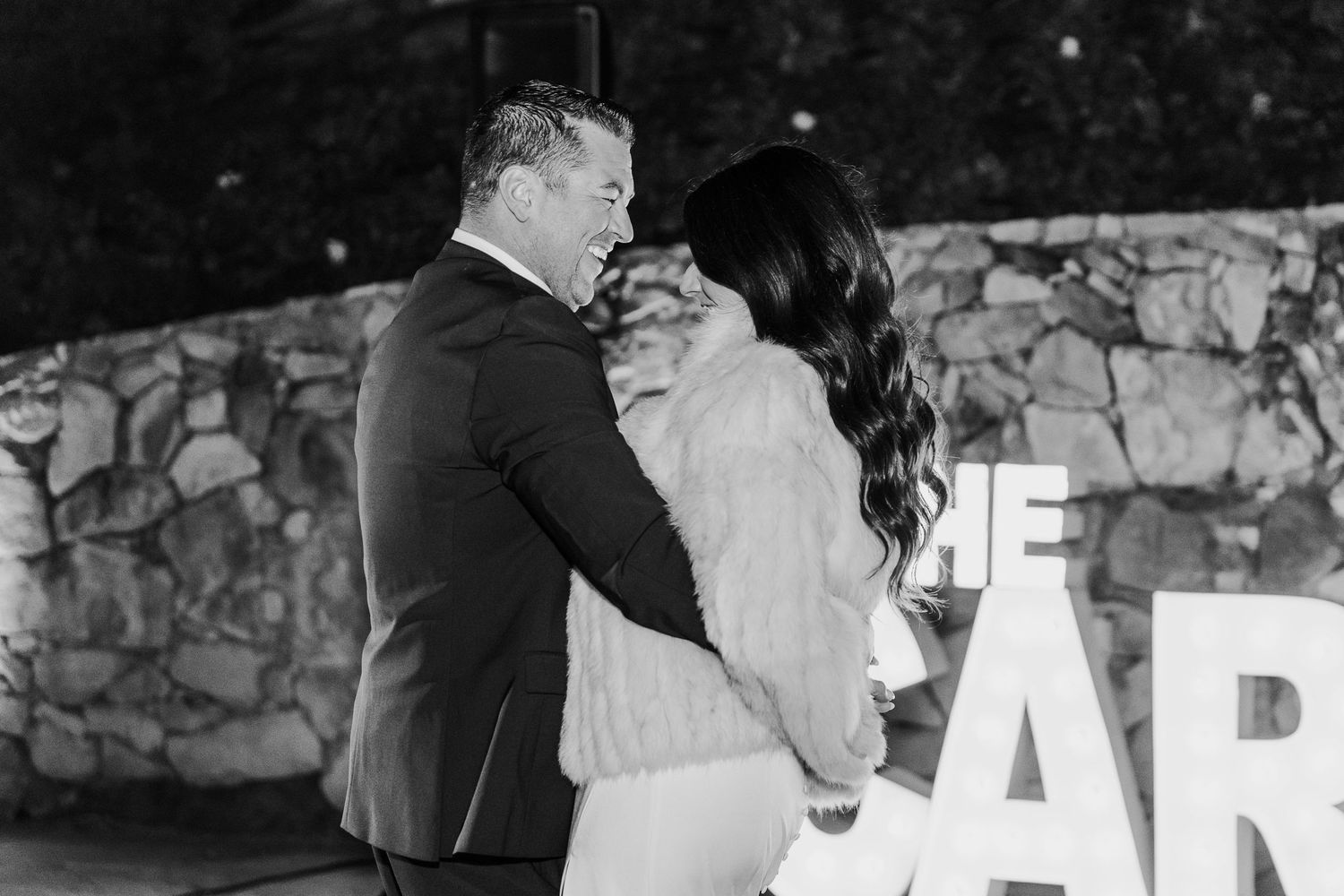 Black and white wedding reception photo with illuminated letter signage against stone wall.