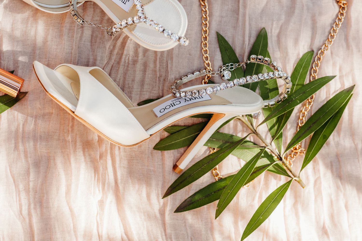 Elegant white bridal heels with crystal embellishments displayed with green leaves on pink silk fabric.