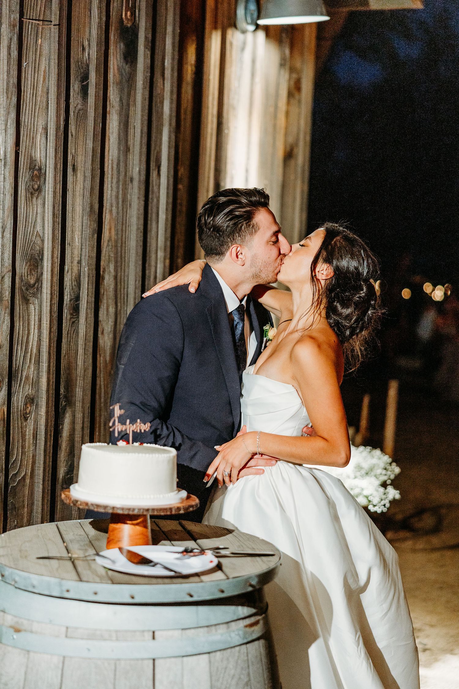Newlyweds sharing cake at their rustic barn wedding reception with wooden barrel table.