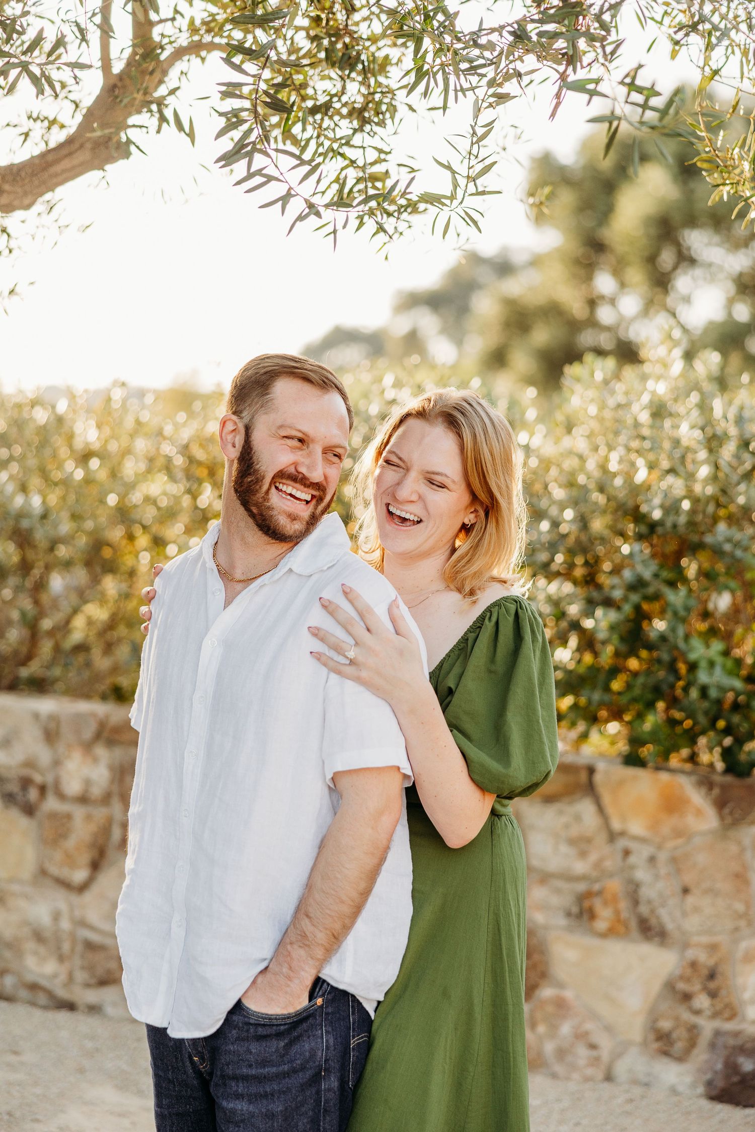 A joyful couple laughs together outdoors against a stone wall backdrop with trees overhead.