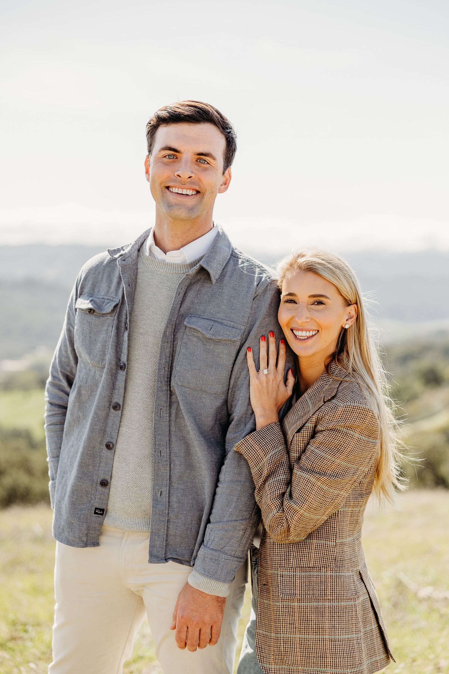 Couple wearing coordinated grey and plaid outfits poses in scenic hillside setting.