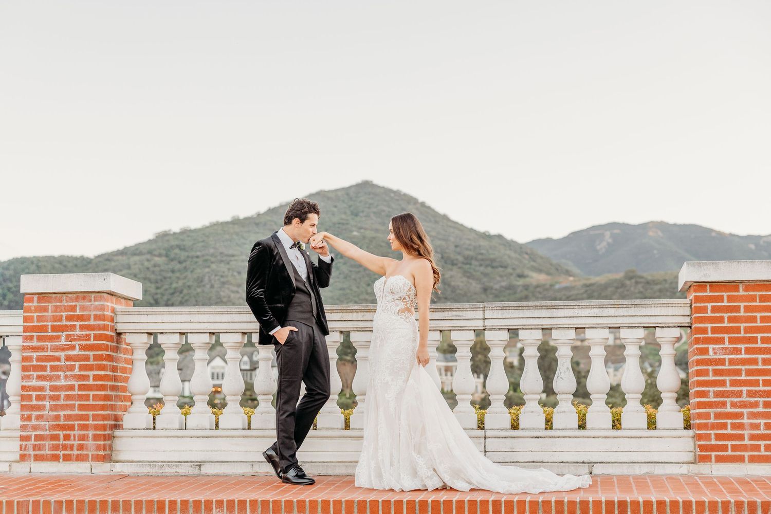 Wedding couple poses along white balustrade with scenic mountain backdrop and brick columns.