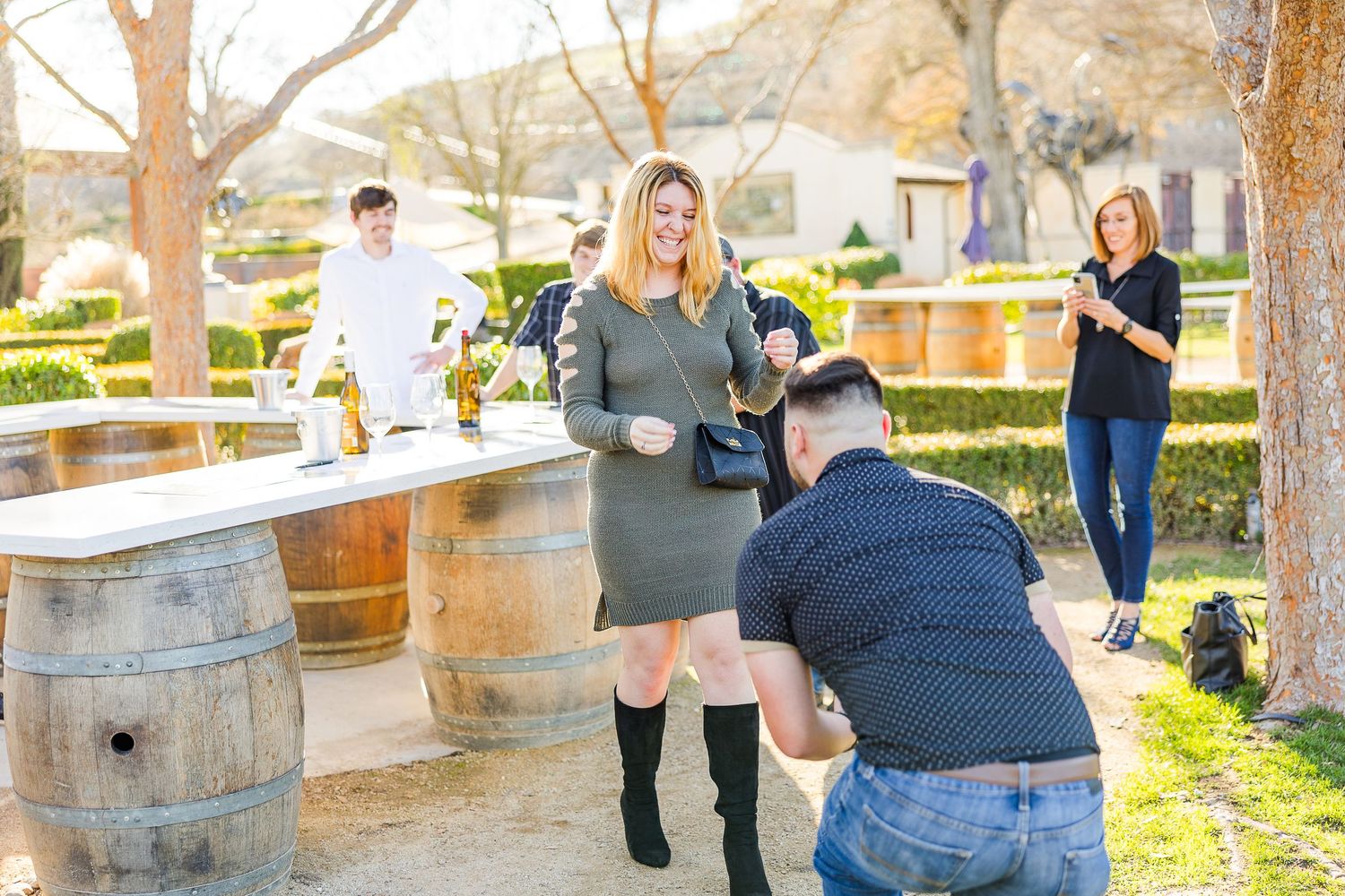 A couple shares a special moment at an outdoor winery with wooden barrels and onlookers in the background.