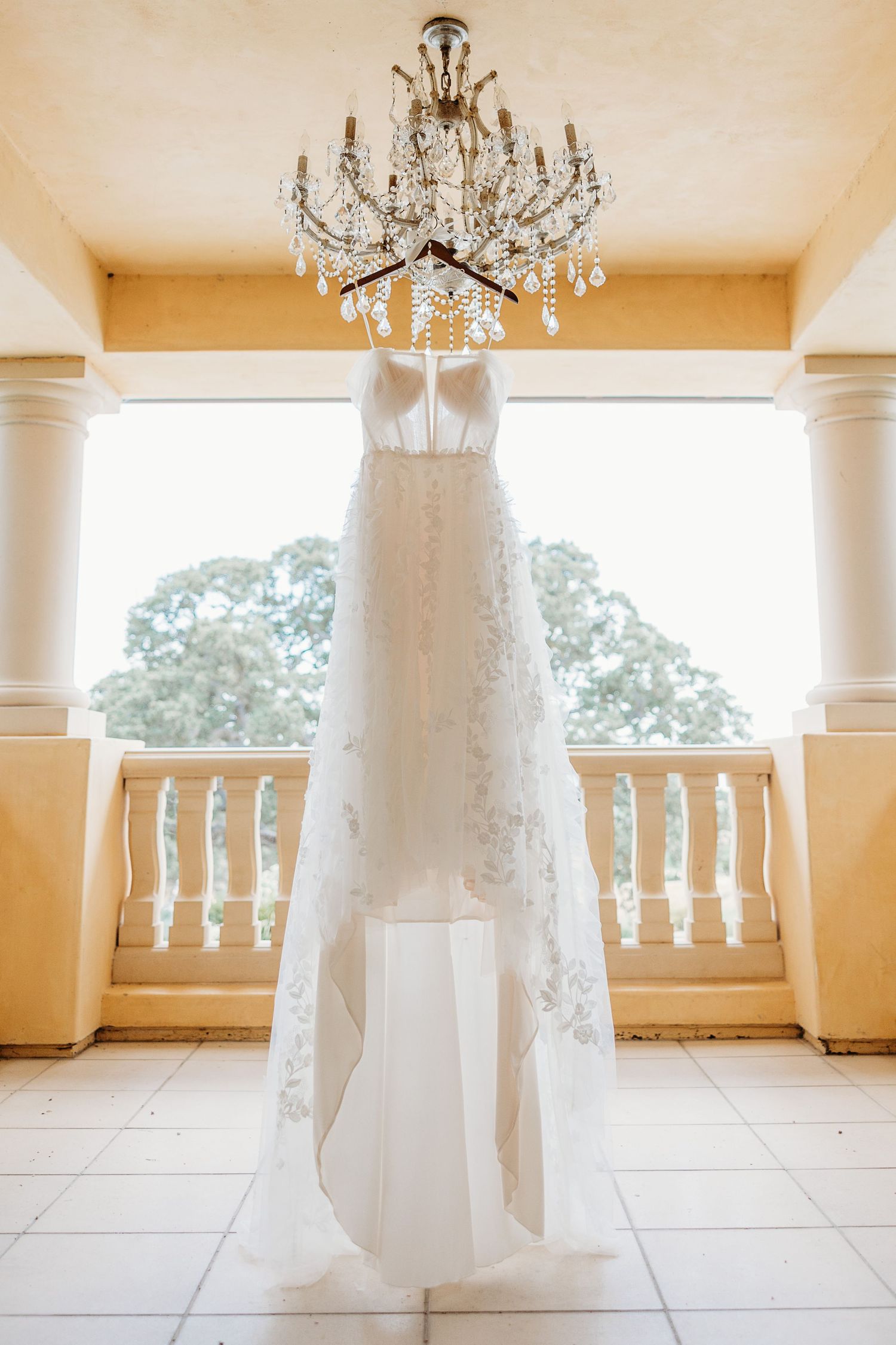White lace wedding dress hanging from crystal chandelier against cream colored balcony.