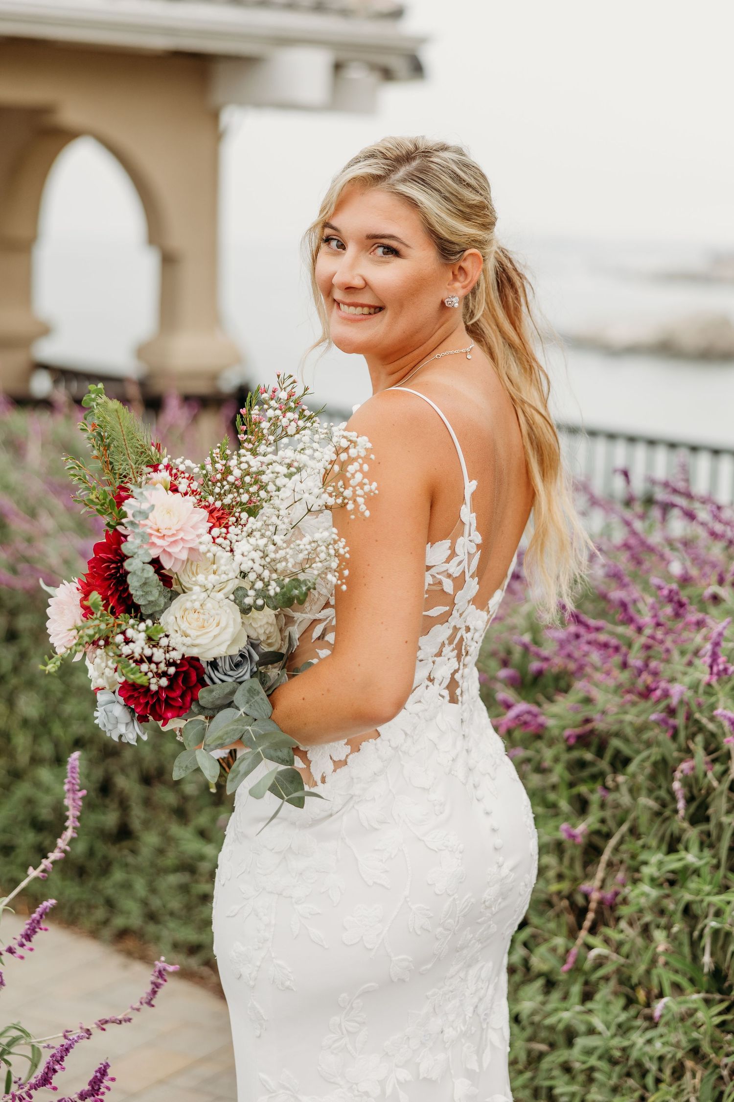 Wedding dress with detailed lace back photographed among coastal gardens with red and white bouquet.