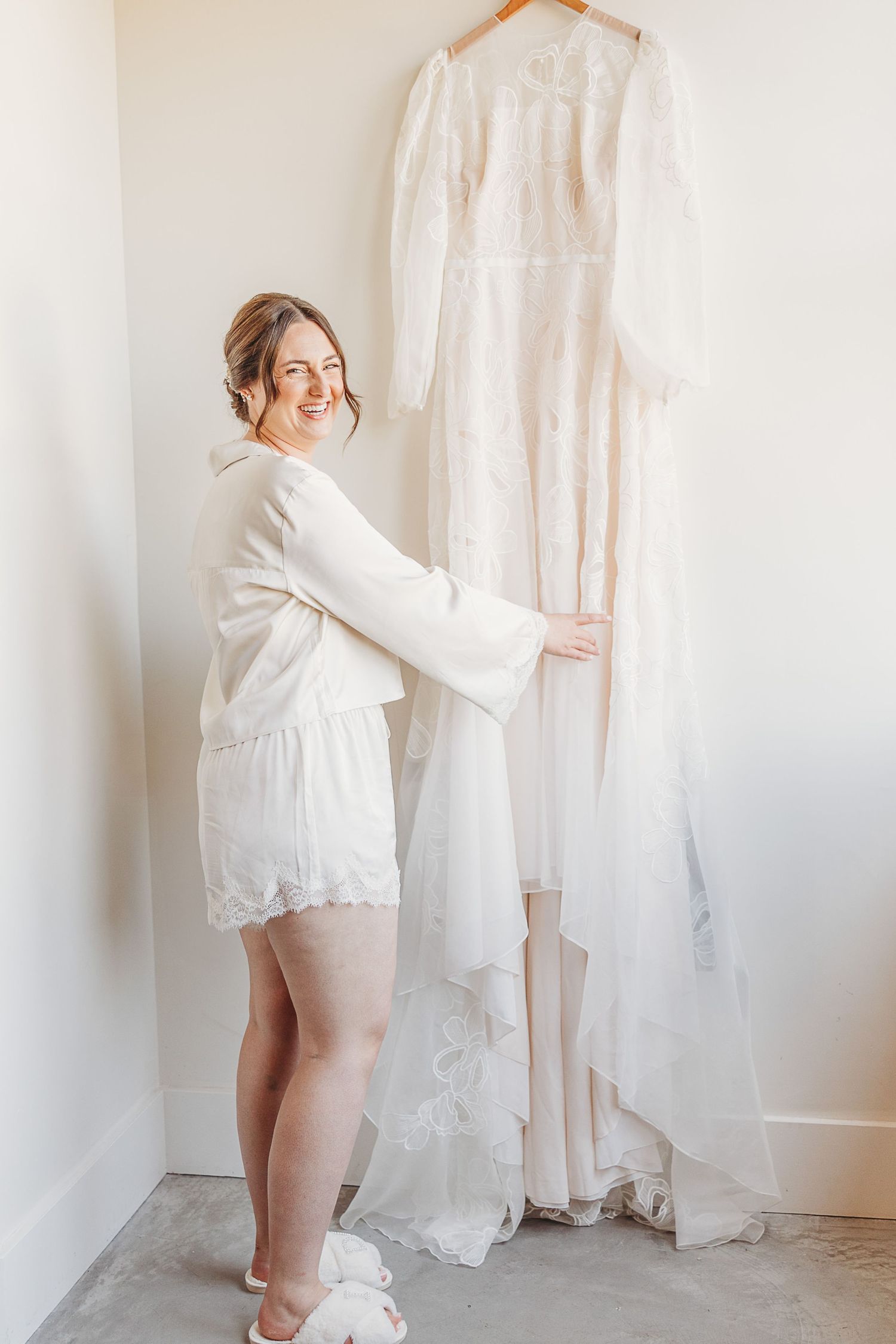 Wedding dress hangs in preparation as someone in white robe poses beside it.