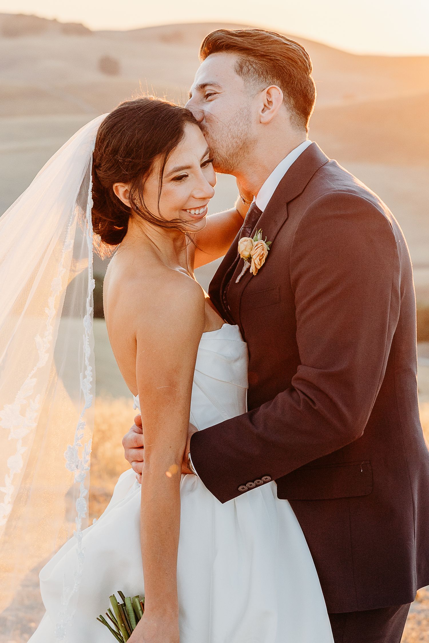 Romantic wedding couple embracing during golden hour sunset with flowing veil.
