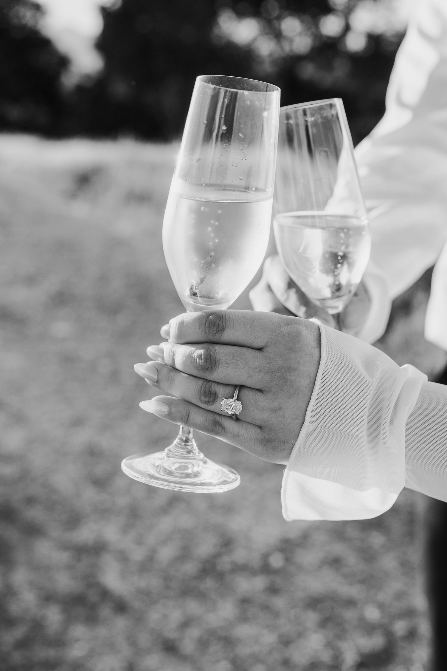 Black and white photo showing hands holding wine glasses in a celebratory toast outdoors.
