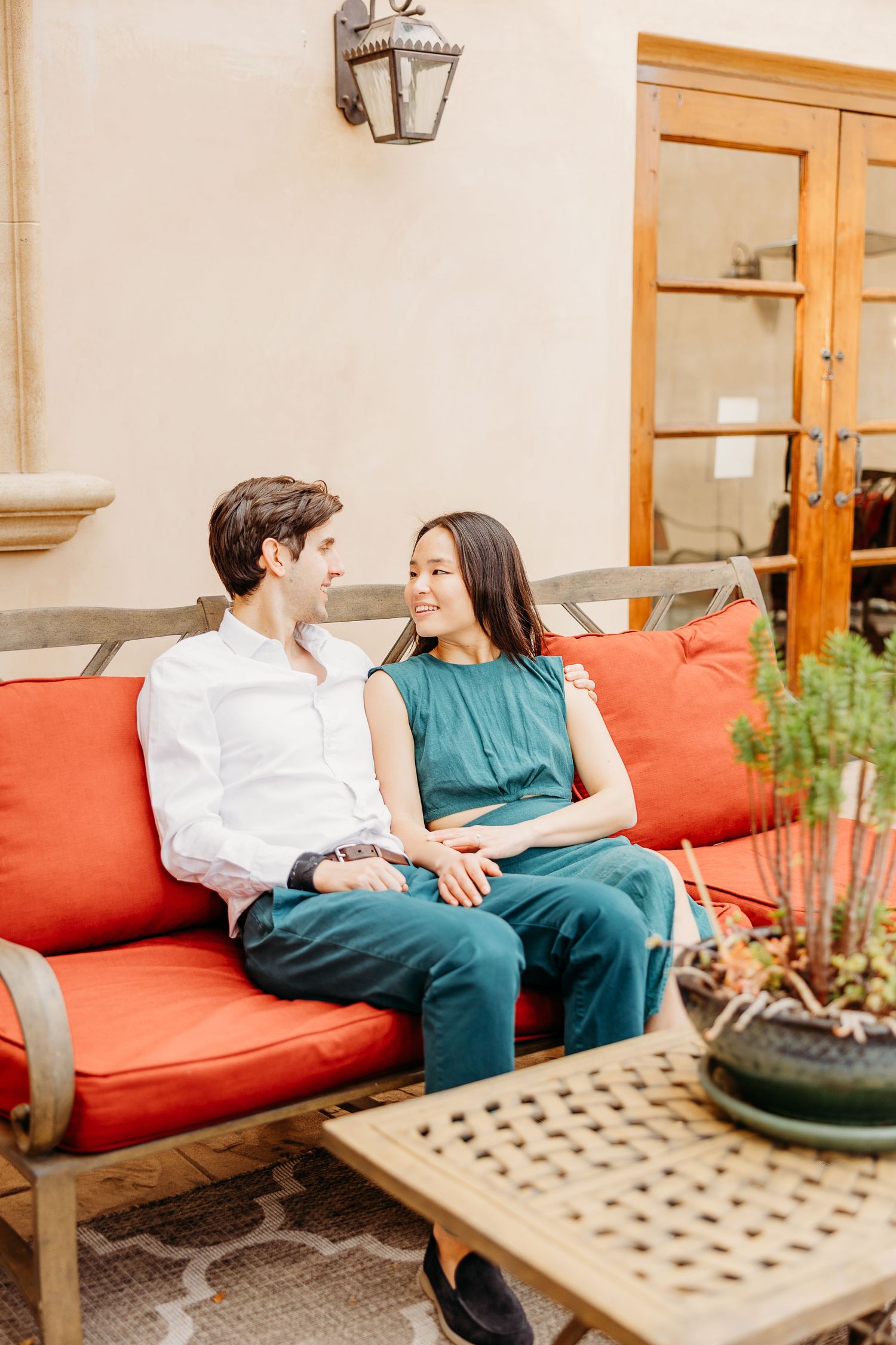 A couple sits intimately on a red cushioned outdoor bench with a decorative wooden table nearby.