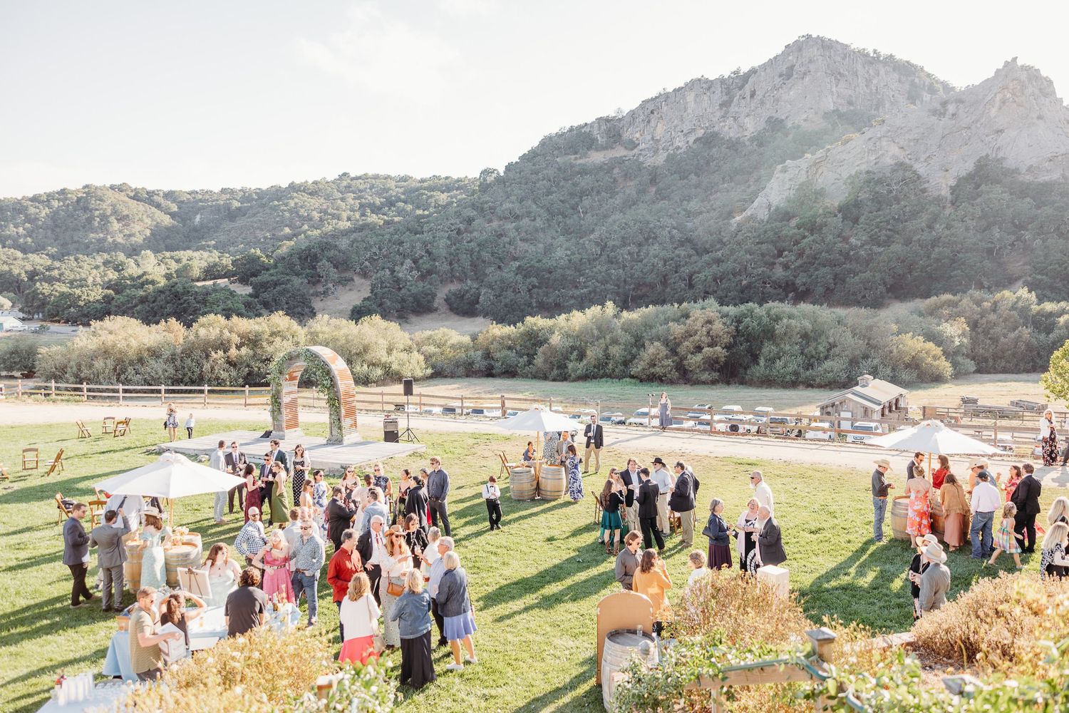 Guests gather at an outdoor wedding ceremony site with mountain views and rustic pastoral scenery in the background.