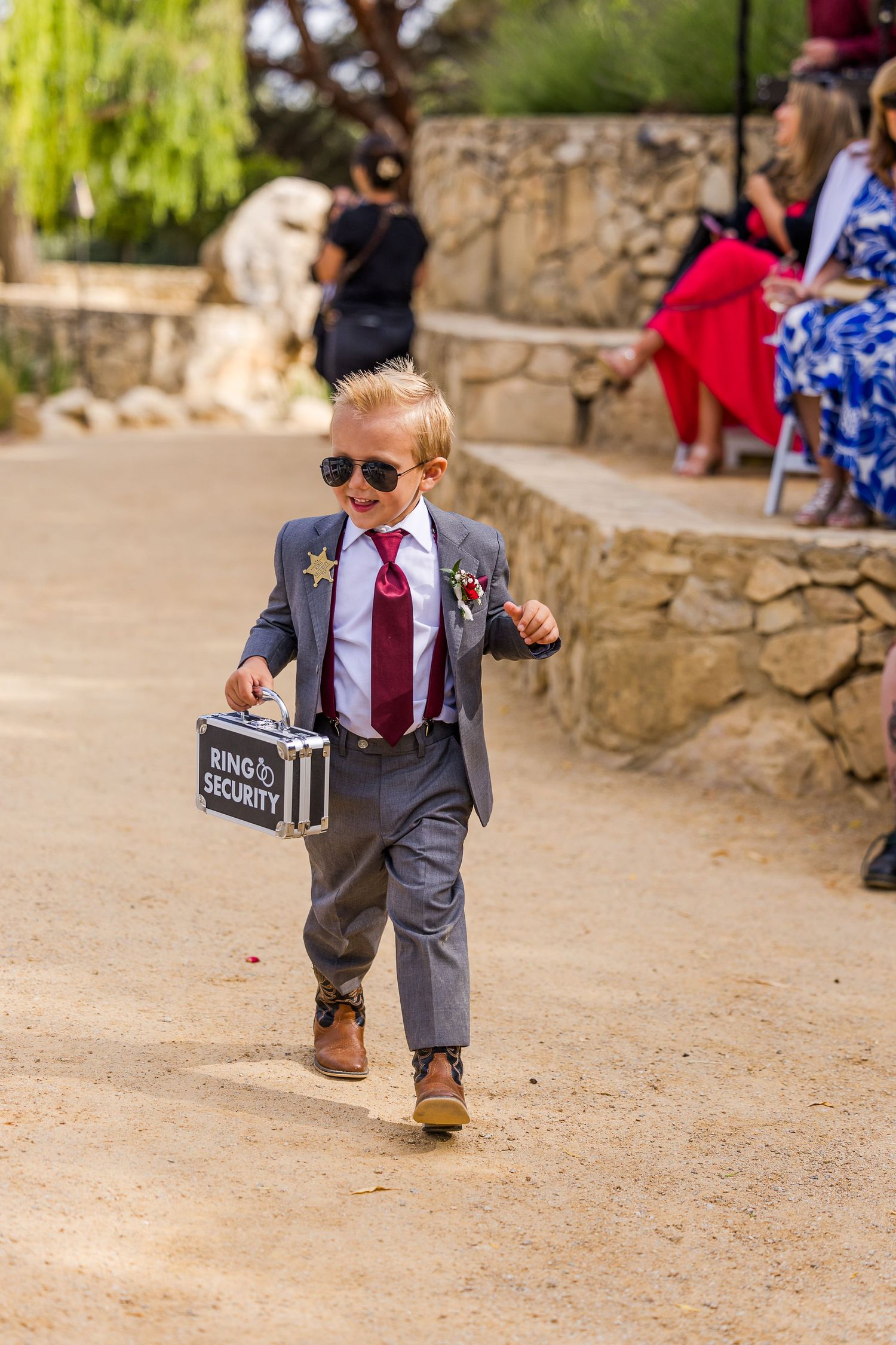 A young ring bearer in a grey suit and sunglasses carries a sign down a stone pathway.