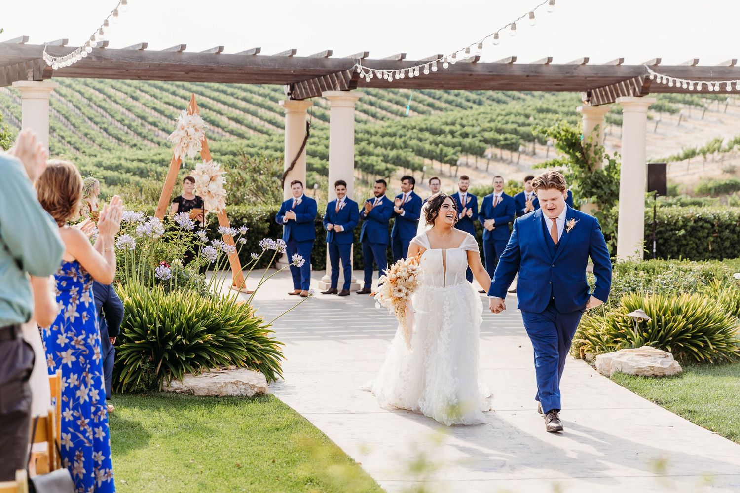 Wedding celebration at outdoor vineyard venue with guests and wedding party in blue attire.