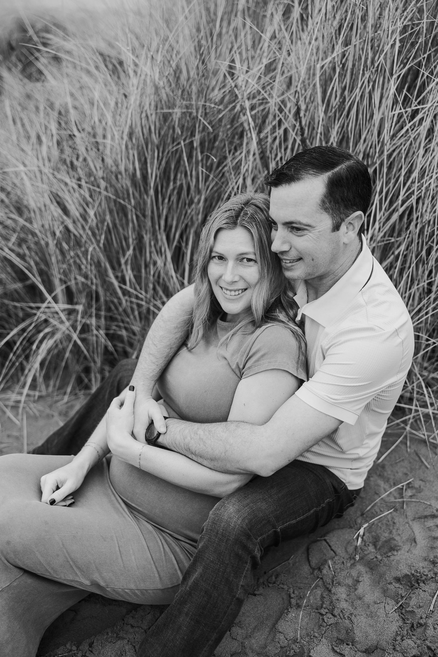 Black and white photo of couple sitting together in tall grass during maternity photo shoot.