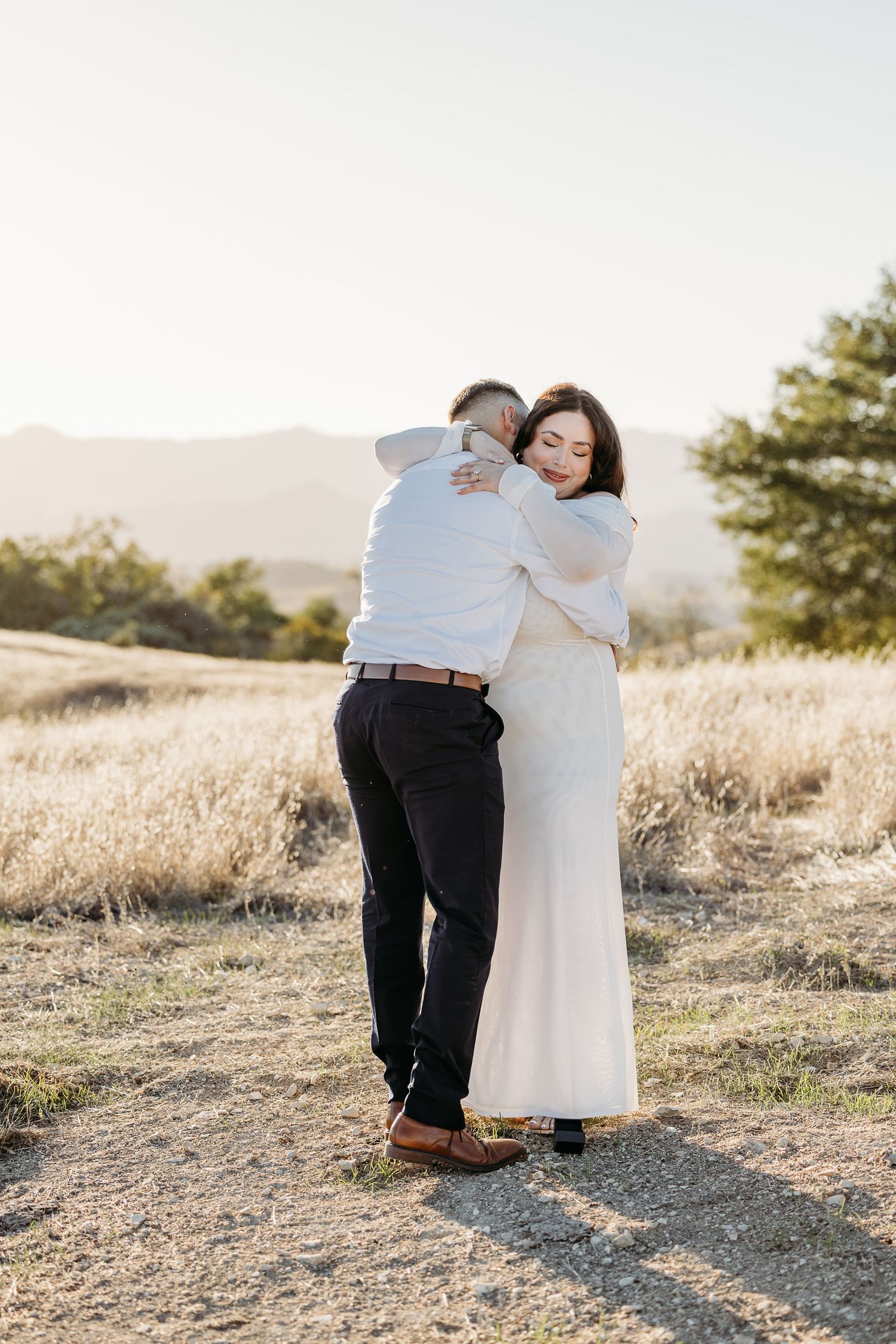 A couple in white and black attire embrace lovingly on a sunlit hilltop during a golden hour photoshoot.