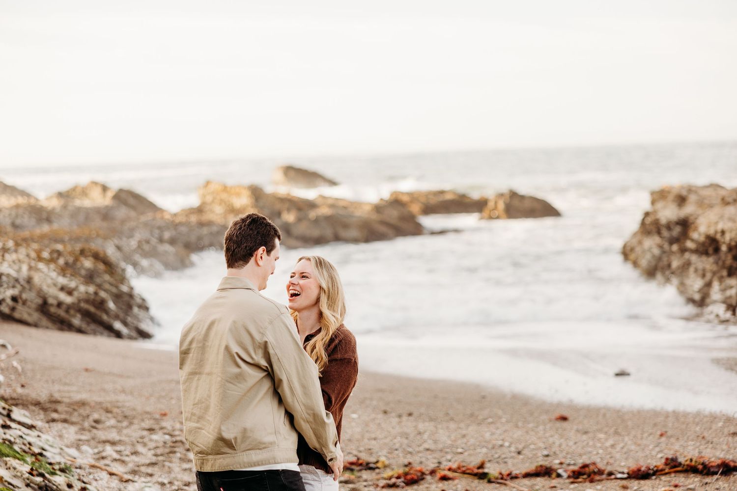 A couple sits together on rocks by the ocean during sunset at a beach in California.