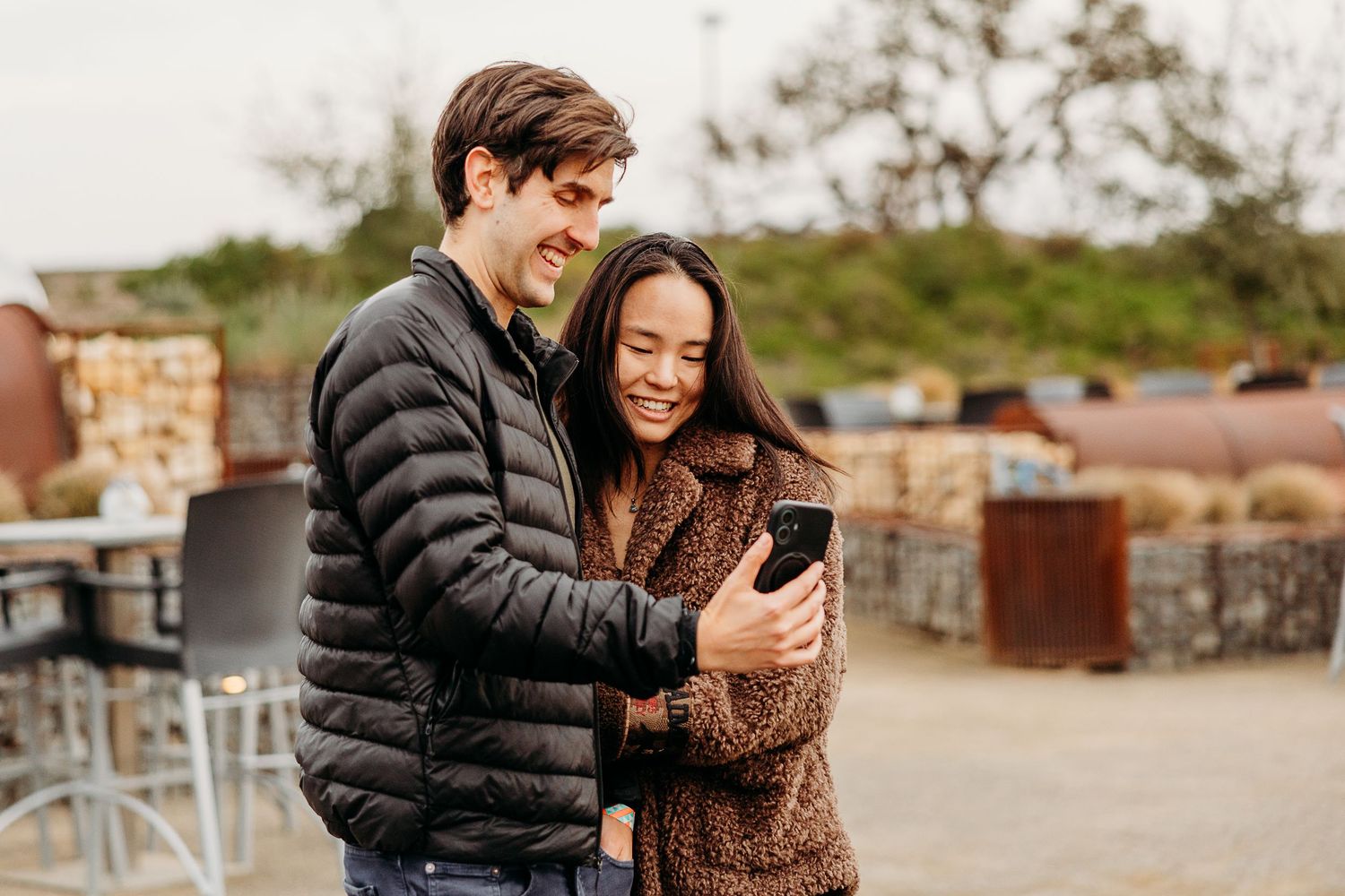 Couple in winter coats looking at mobile phone while standing together outdoors on autumn day.