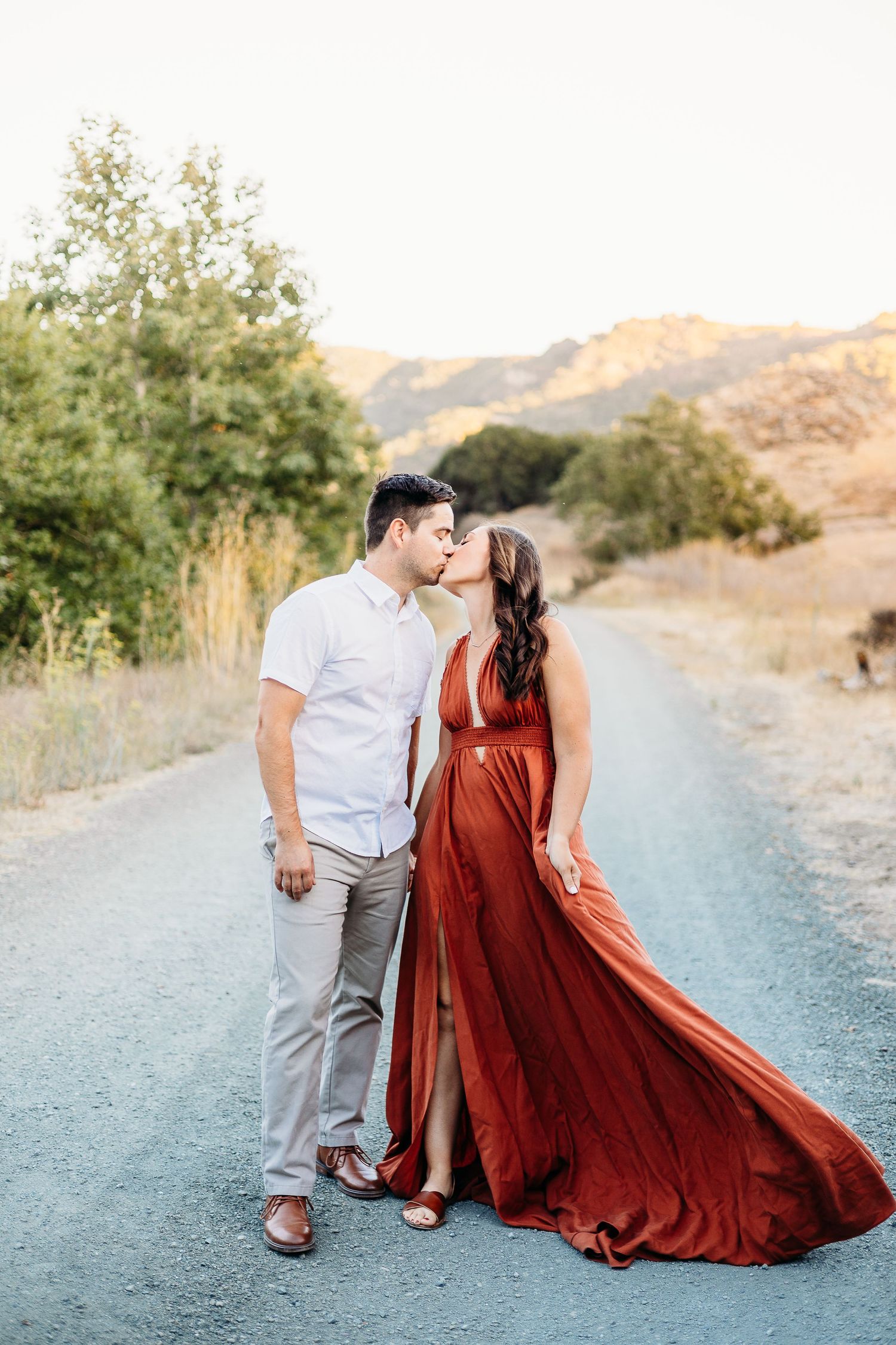 A romantic couple shares a tender moment on a dirt path with mountains in the background.