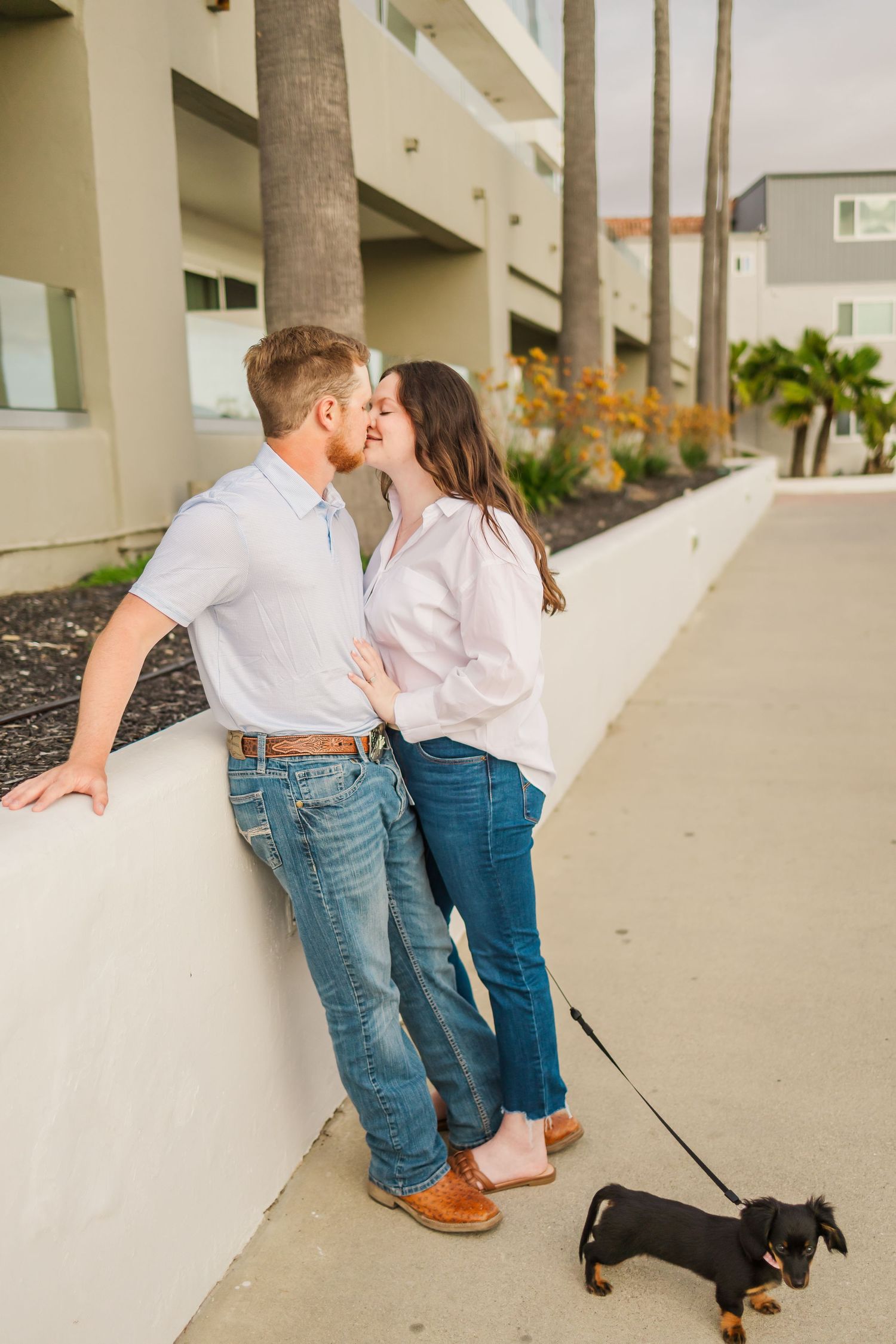 Romantic couple sharing a kiss against white wall while their dachshund looks on.